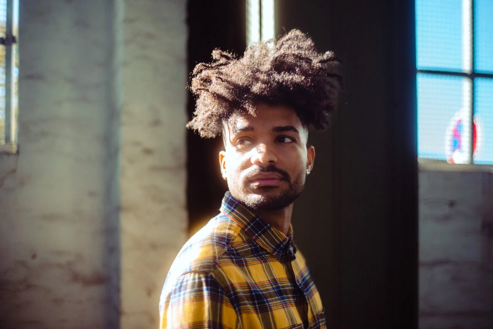 Junger Mann mit lockigem Haar in einem gelb-gestreiften Hemd steht vor einer Wand mit Fenstern und Sonnenlicht, das auf sein Gesicht scheint.