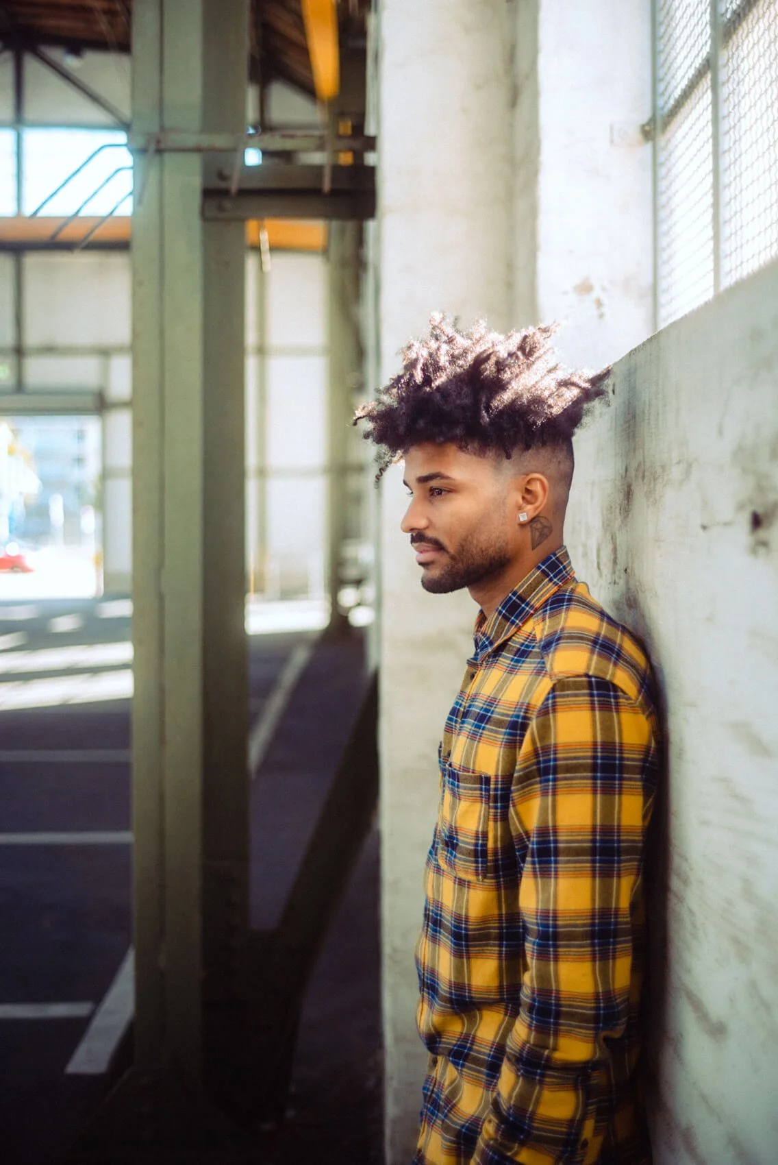 Junger Mann mit dunklem Haar und Vollbart lehnt an einer weißen Wand, trägt ein gelb-blau kariertes Hemd, im Hintergrund eine Turnhalle mit Parkplätzen und Metalltreppe.