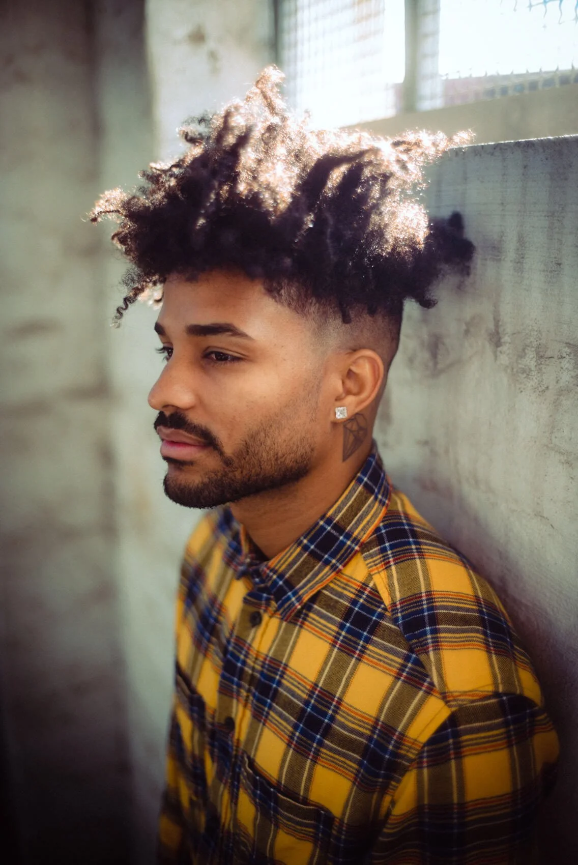Junge mit vollem, lockigem Haar, tätowiertem Hals, Earring und kariertem Hemd, lehnt an einer Wand in einem Industrie- oder Lagerraum.