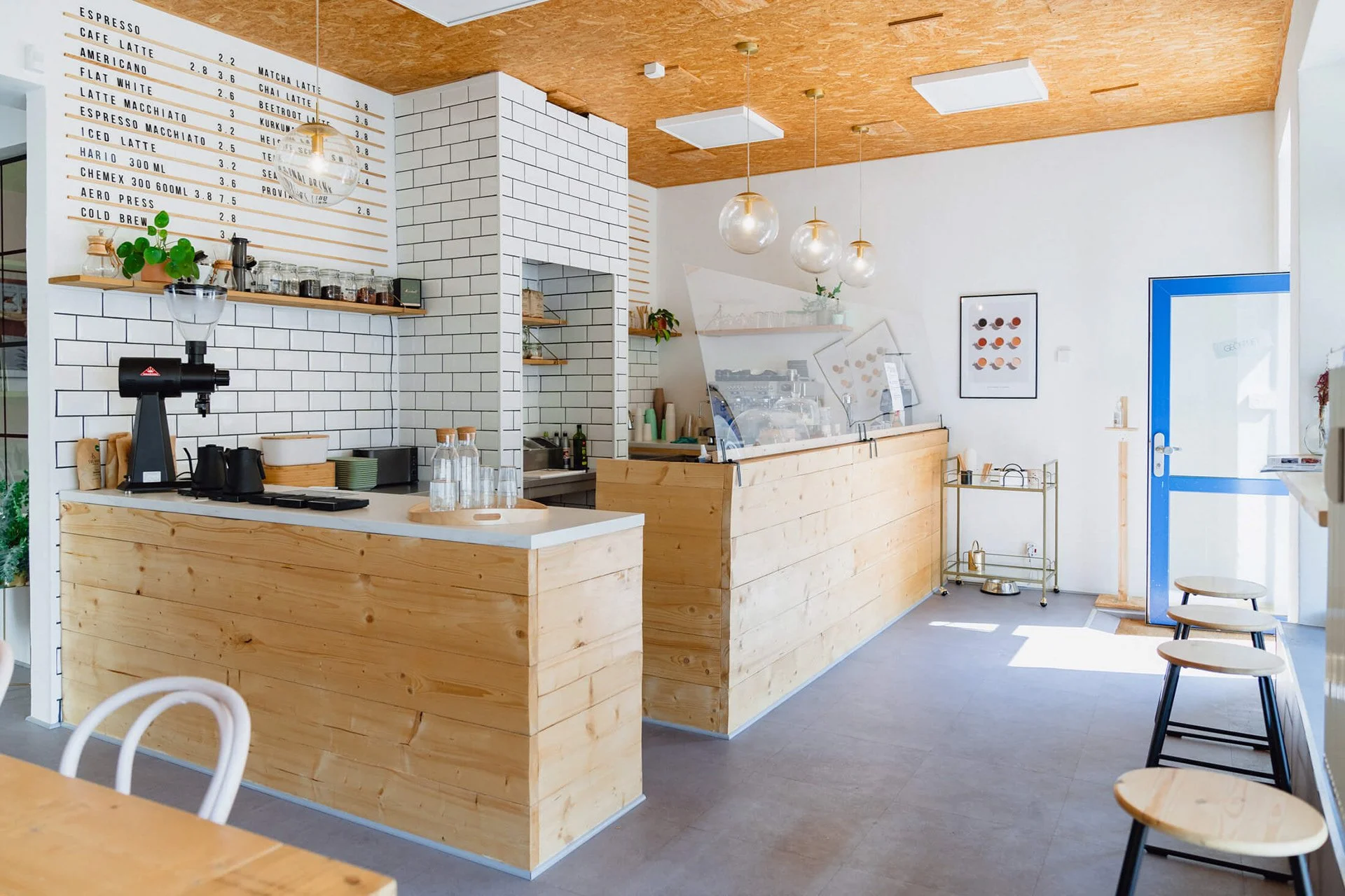 Ein modernes Café mit Holztheke, weißer Wand mit schwarzer Menütafel, steinernen Akzenten, dekorativen Lampen und Sitzgelegenheiten aus Holz und Metall.