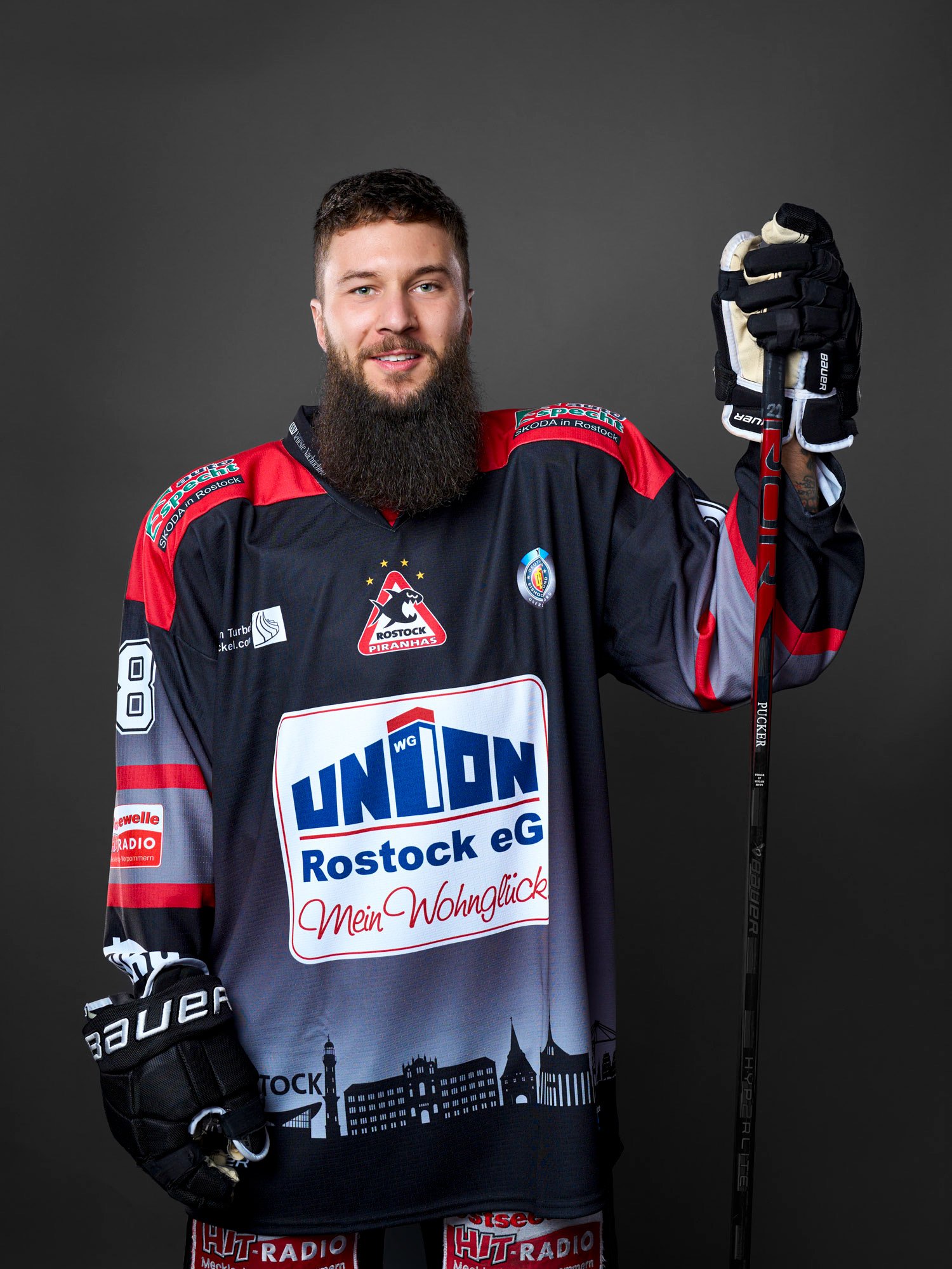 Eishockeyspieler der Rostock Piranhas mit dunklem Trikot und Rucksack, hält Eishockeyschläger in die Höhe, steht vor grauem Hintergrund.
