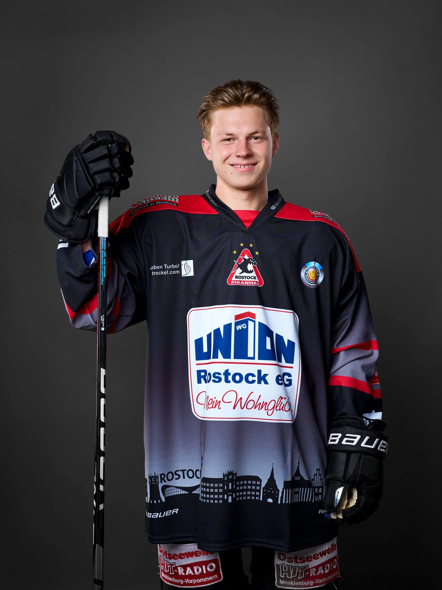 Junger Eishockeyspieler der Rostock Piranhas in dunklem Trikot mit rotem und grauem Akzent, hält Eishockeyschläger, trägt Handschuhe und lächelt vor dunklem Hintergrund.