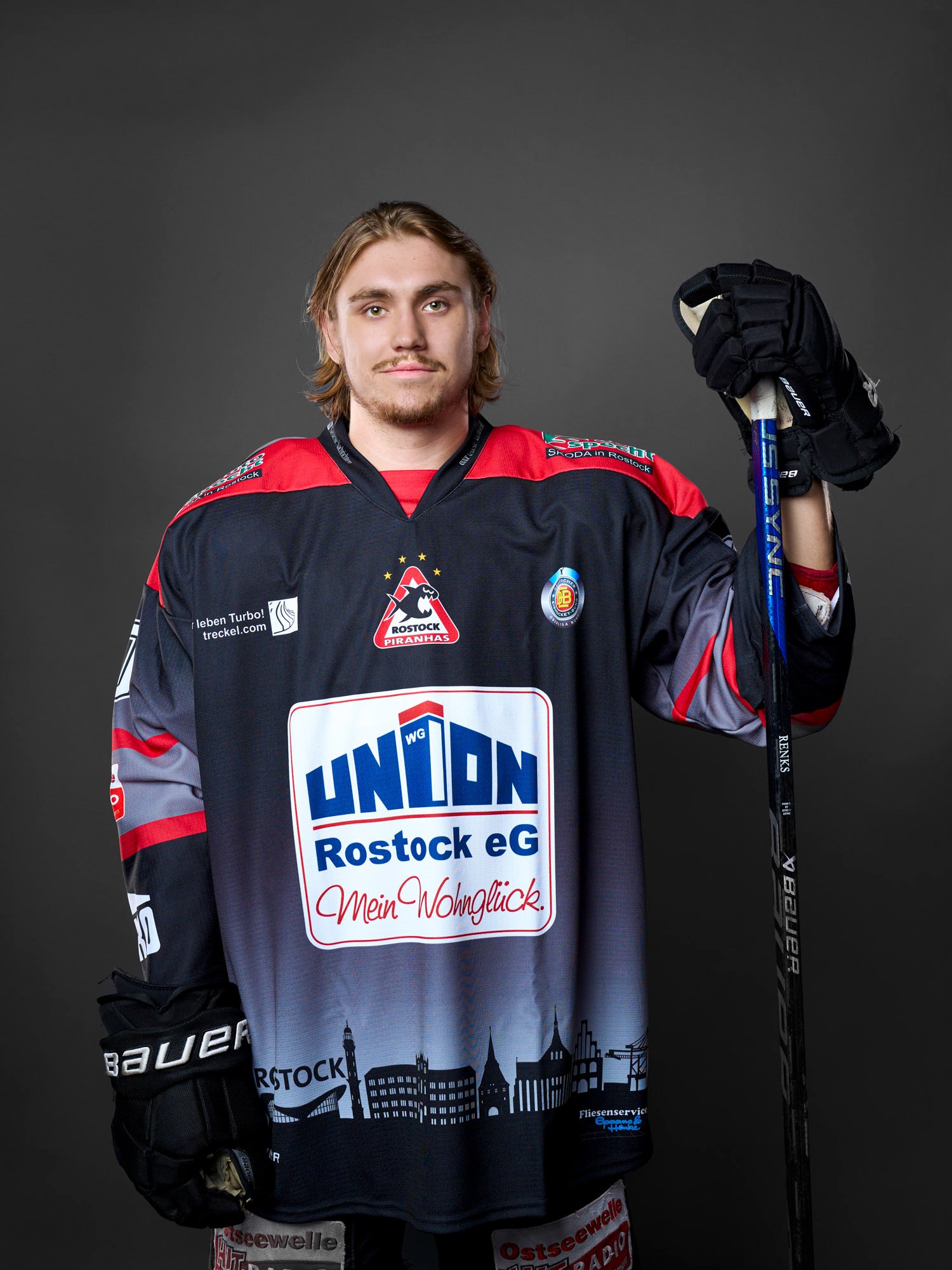 Junger Eishockeyspieler der Rostock Piranhas in schwarzer, roter und grauer Eishockeyausrüstung, der ein Hockeyschläger in der Hand hält, vor einem dunklen Hintergrund.