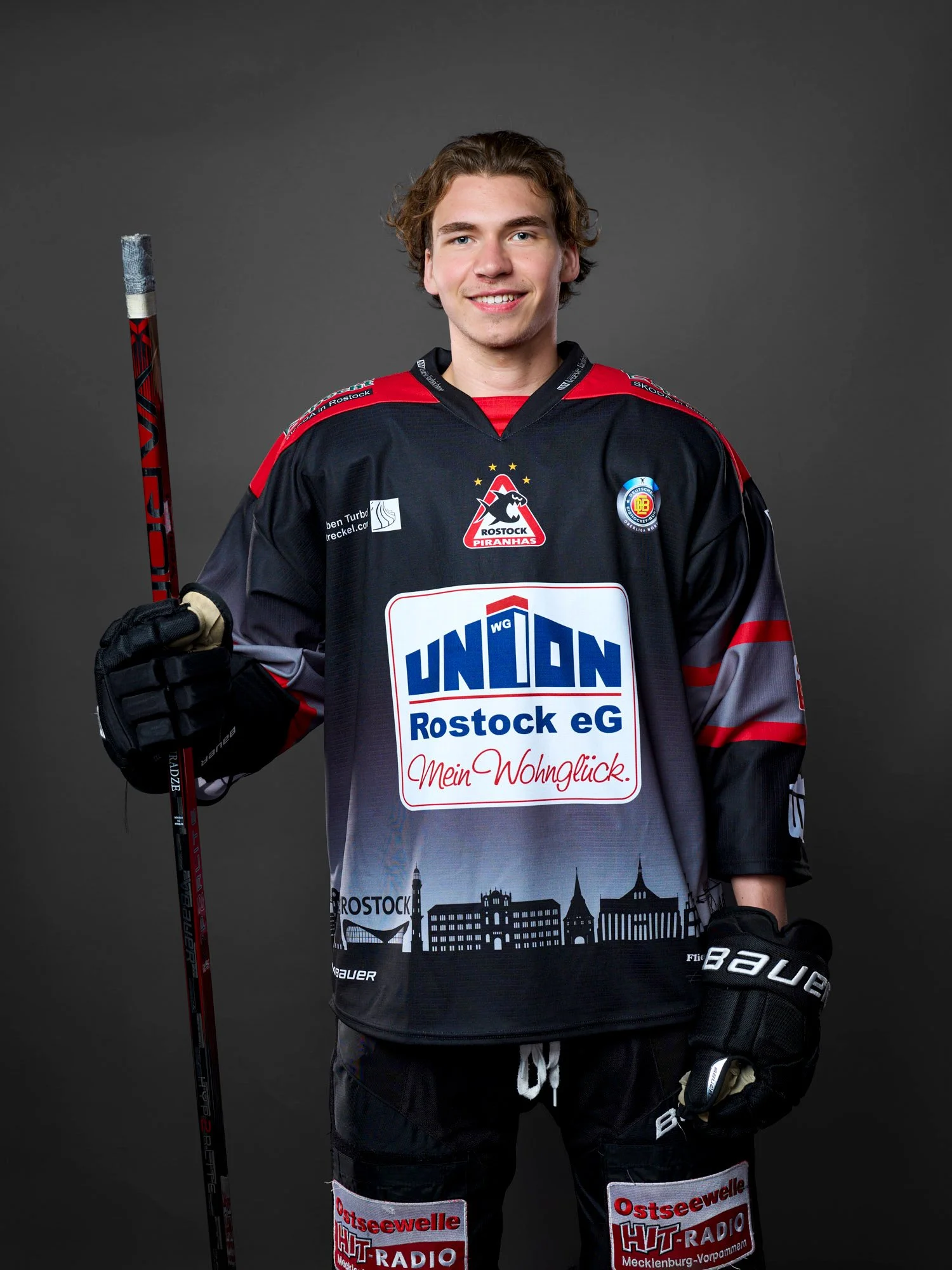 Junger Eishockeyspieler der Rostock Piranhas in schwarzer Mannschaftsuniform mit mehreren Logos, hält einen Eishockeyschläger, vor dunklem Hintergrund.