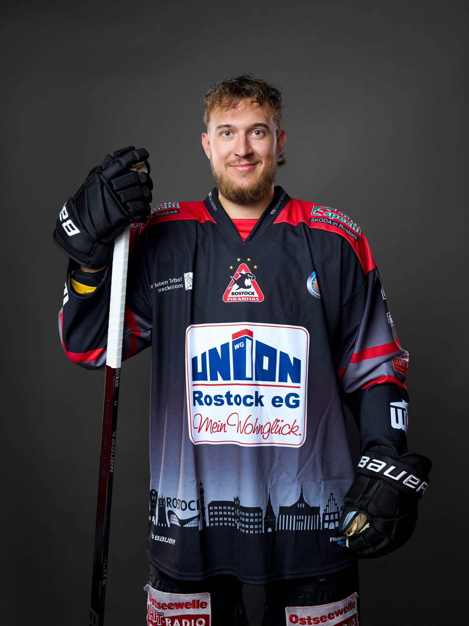 Ein junger Mann in Eishockeytrikot der Rostock Piranhas mit Eishockeyschläger, der auf einem grauen Hintergrund posiert.