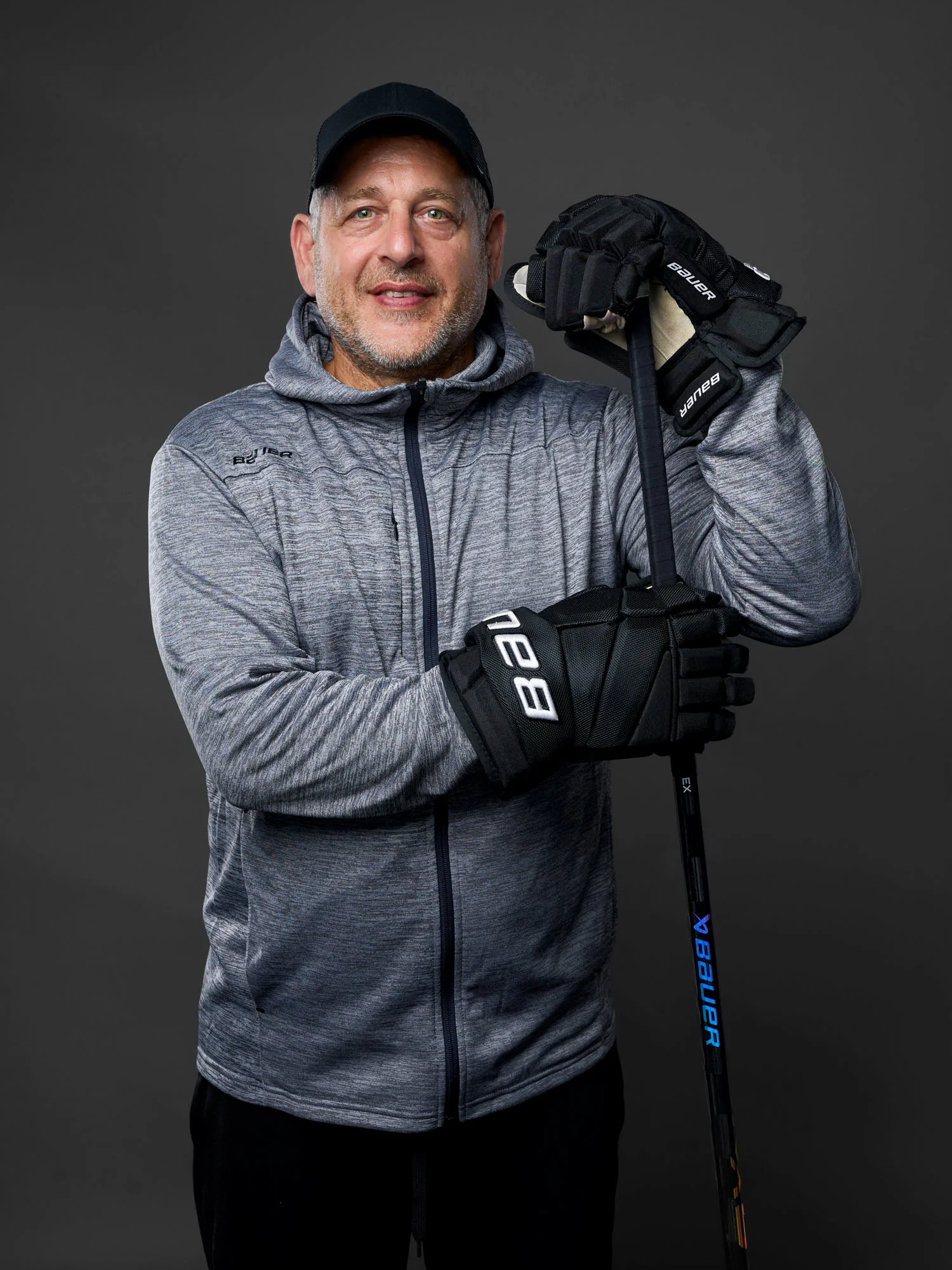 Der Trainer der Rostock Piranhas trägt einen grauen Sportpullover, eine schwarze Mütze und Hockeyhandschuhe und hält einen Eishockeyschläger. Er steht vor einem grauen Hintergrund.