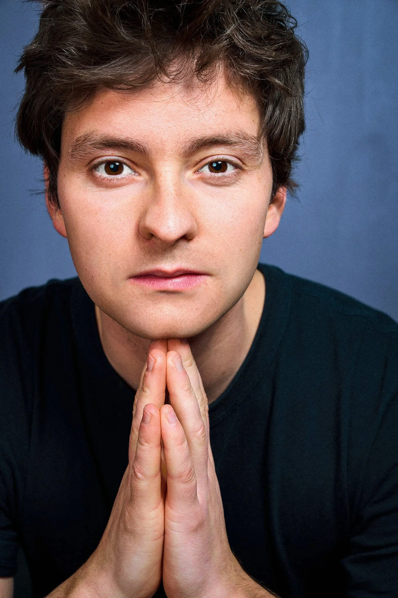 Daniel Littau mit dunklem Haar, in Schwarz gekleidet, mit zusammengelegten Händen vor seinem Kinn, schaut ernst in die Kamera, blauer Hintergrund.