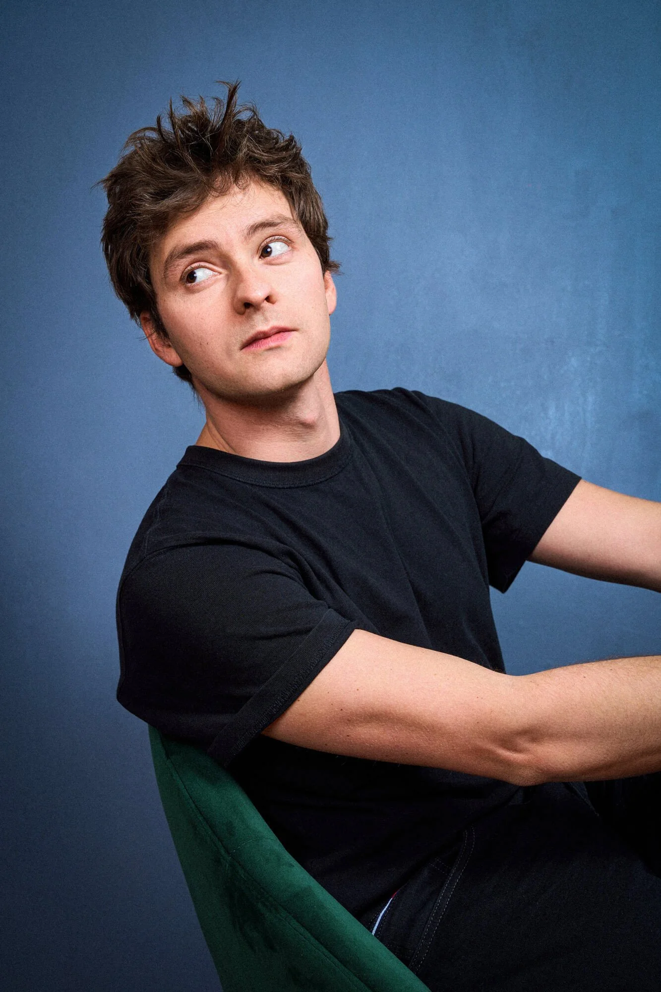 Daniel Littau sitzt in einem schwarzen T-Shirt auf einem grünen Stuhl vor einem blauen Hintergrund, schaut nach oben und zur Seite.