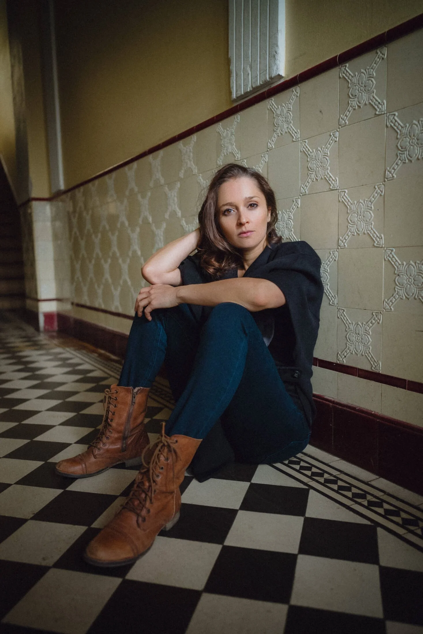 Sarah Alles sitzt auf dem Boden eines Flurs, trägt braune Stiefel, Jeans und eine schwarze Jacke. Sie schaut direkt in die Kamera.