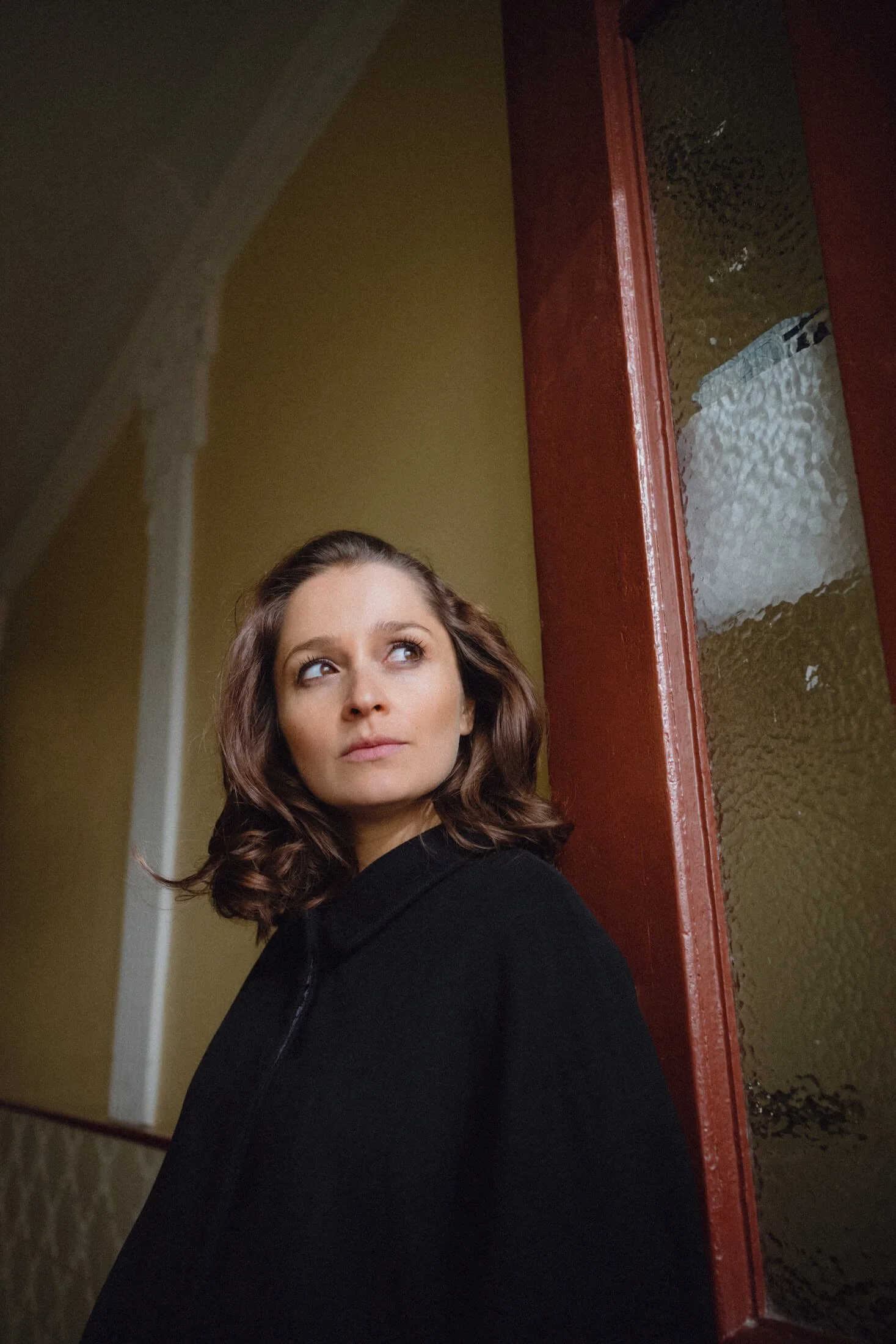 Portrait von Schauspielerin Sarah Alles mit braunen, lockigen Haaren, die gegen eine rote Tür und eine gelbe Wand lehnt, blickt nach oben.