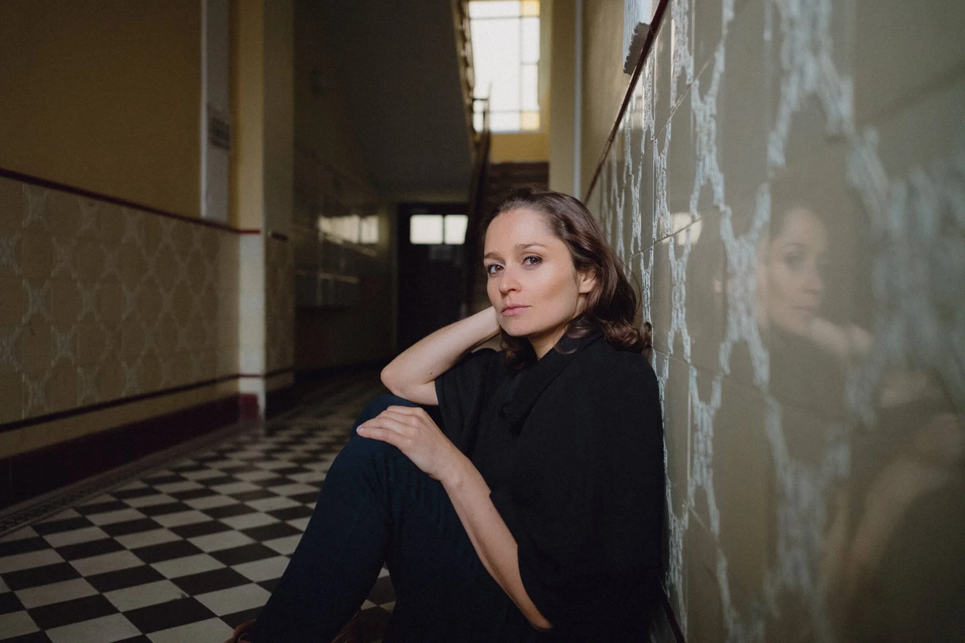 Sarah Alles sitzt an einer Wand in einem Flur, lehnt mit dem Rücken an die Wand und schaut nach vorne, die Wand ist mit Fliesen bedeckt, ihr Spiegelbild ist auf der Wand zu sehen.