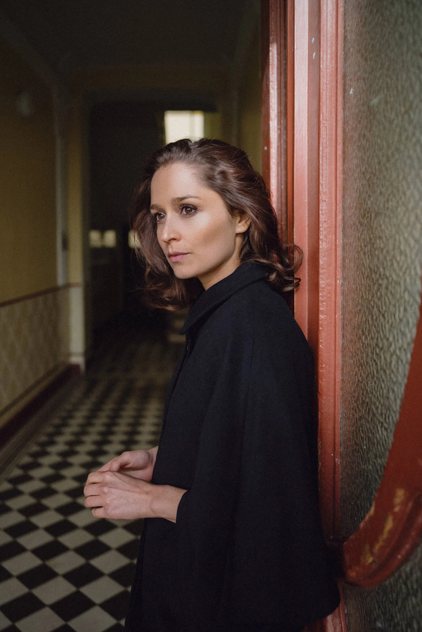 Sarah Alles mit braunen, gewellten Haaren, die an einer Wand in einem Flur lehnt, mit einem nachdenklichen Blick.