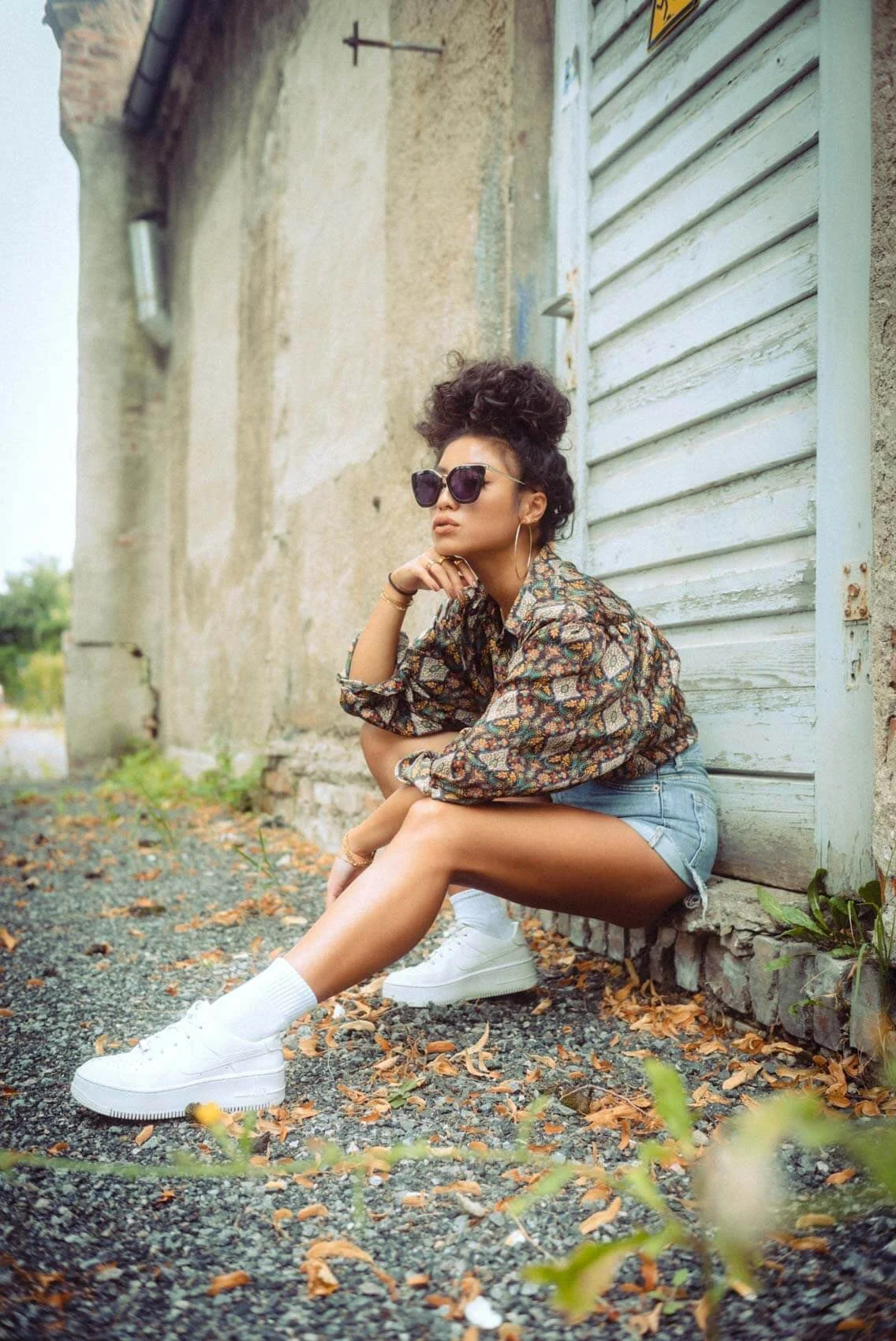 Junge Frau mit großen Sonnenbrillen, lockigem Haar, kariertem Hemd, Shorts und Turnschuhen, sitzt auf dem Boden vor einer alten Steinmauer und einer Holzgarage, umgeben von Herbstblättern.