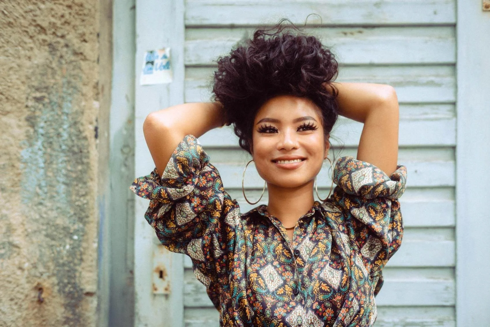 Eine junge Frau mit dunklen, lockigen Haaren, die einen bunten, mit Blumenmuster versehenen Oberteil trägt, lächelt und die Arme hinter dem Kopf verschränkt vor einer alten, holzverkleideten Wand.
