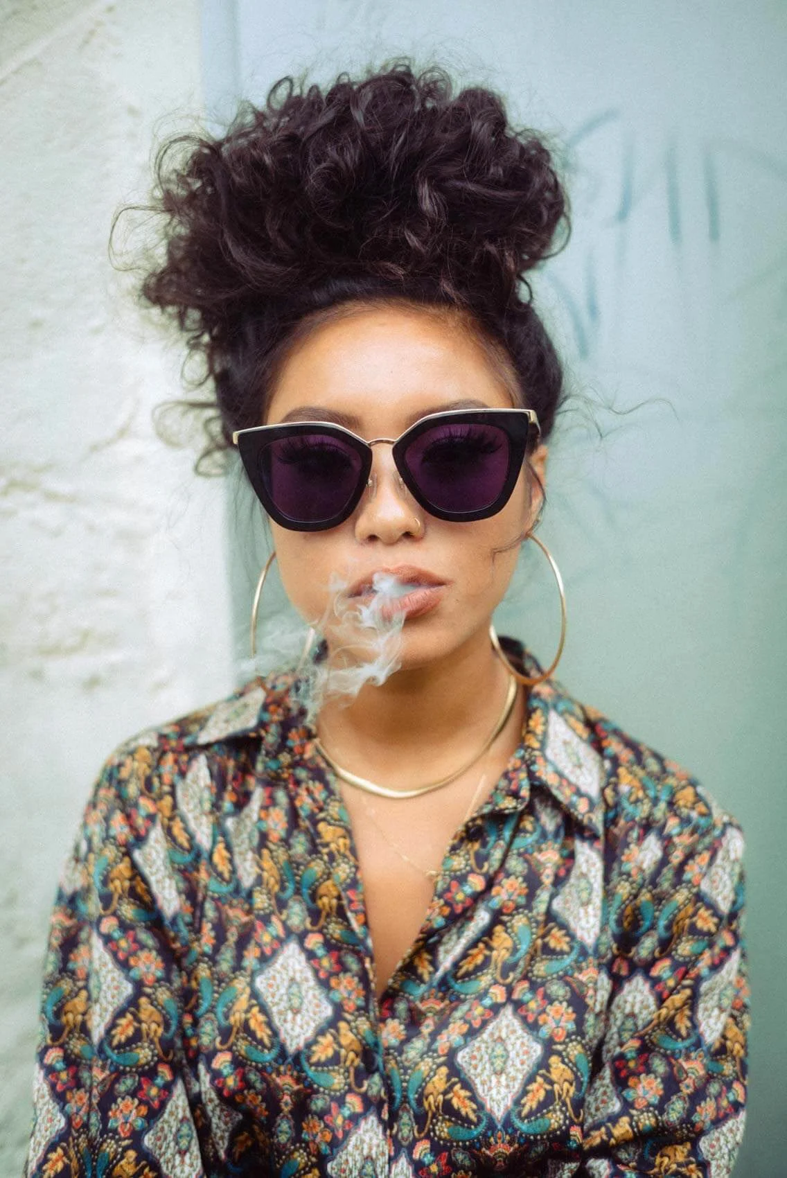 Junge Frau mit großen dunklen Sonnenbrillen, curly Haaren auf dem Kopf, Ohrringen, einem Nasenpiercing, einer Goldkette und einem bunten, gemusterten Hemd, die an der Wand steht und Rauch raucht.
