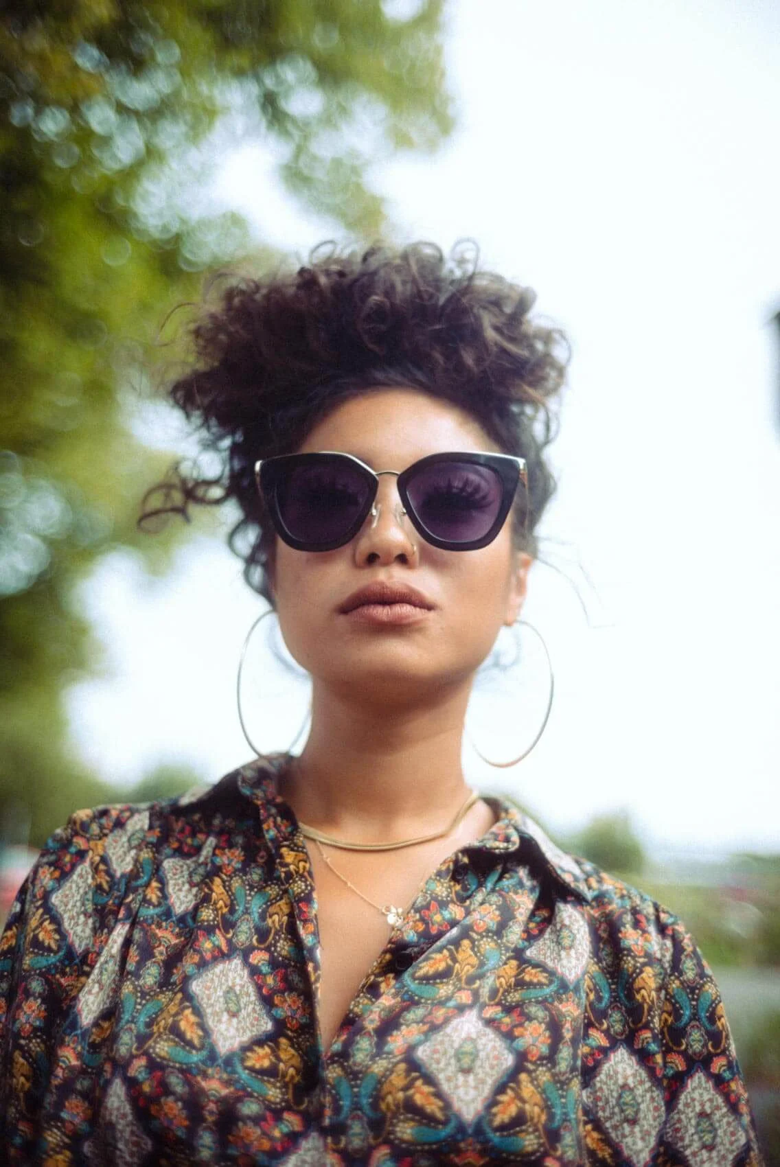 Junge Frau mit dunklen, runden Sonnenbrillen, großen Ohrringen, einem Messinghalsreif und einem bunten, gemusterten Hemd, steht draußen mit Hintergrund aus grünen Bäumen.