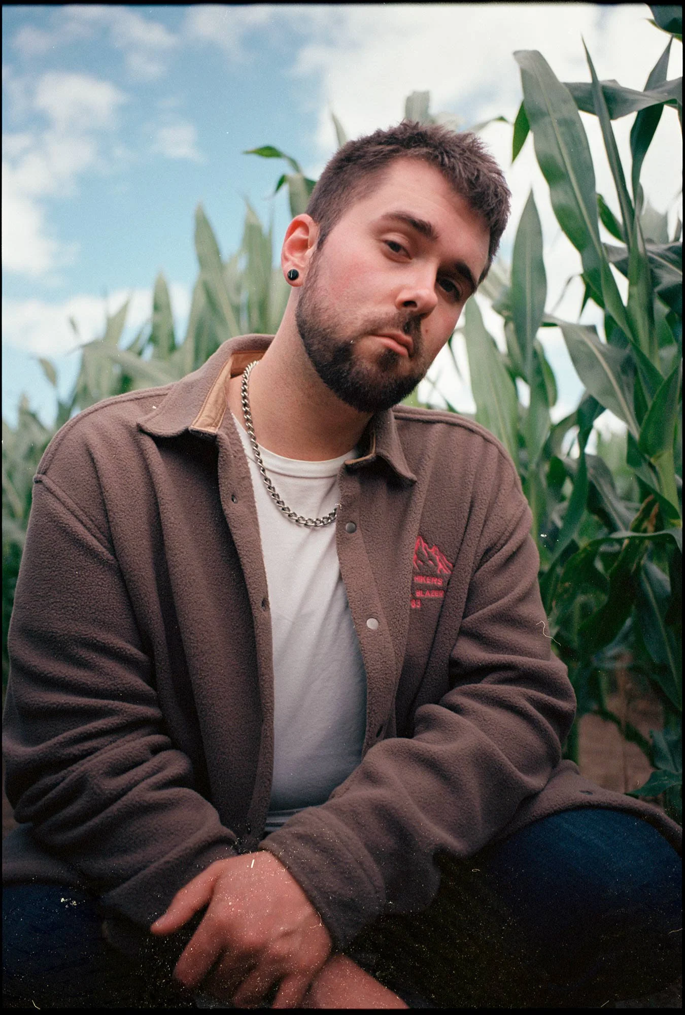 VAUH sitzt in einem Feld mit Maispflanzen, trägt ein braunes Fleeceshirt, eine weiße T-Shirt und eine Silberkette, hat kurze braune Haare, einen Bart und trägt einen Ohrstecker.