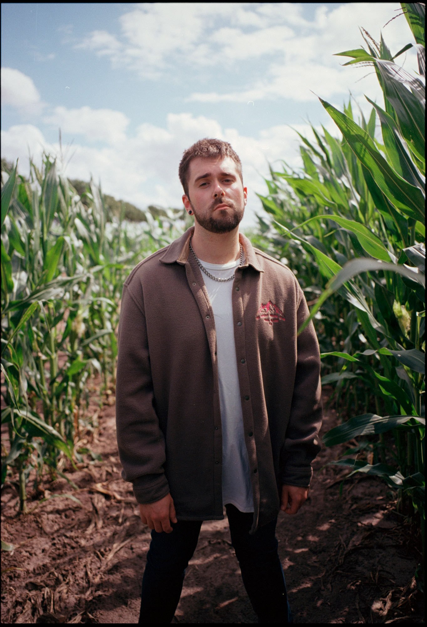 VAUH mit kurzem Haar, Bart und Piercing steht in einer Maisfeld unter einem blauen Himmel mit Wolken.