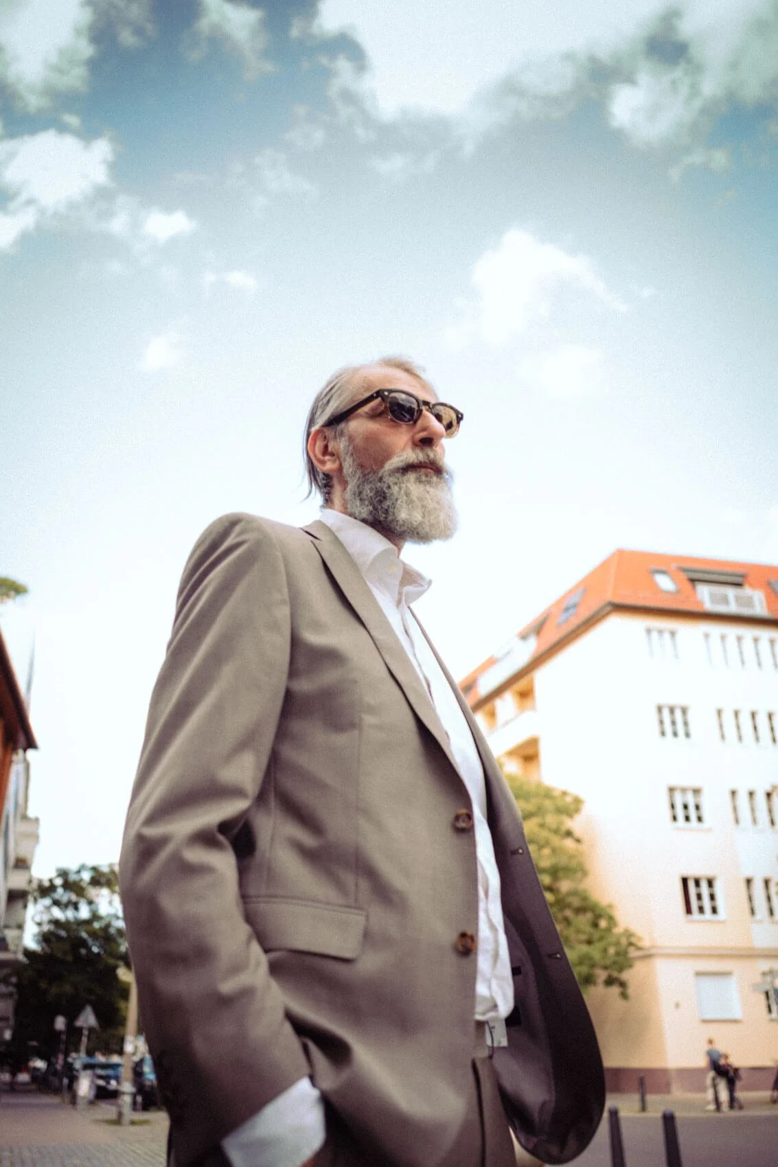 Harald Schindel mit grauem Bart und Brille trägt einen beigen Anzug und steht vor einem mehrstöckigen Gebäude mit roten Dächern, während er in den Himmel blickt.
