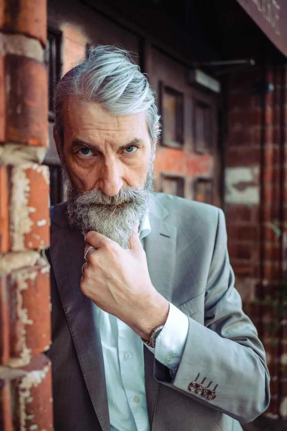 älterer Mann mit grauem Bart und Haaren in einem grauen Anzug, der nachdenklich wirkt, an einer roten Backsteinmauer stehend.
