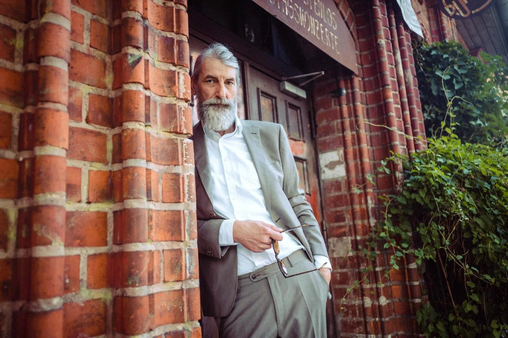 Harald Schindel in einem grauen Anzug lehnt an einer roten Ziegelmauer vor einem Holzschild.