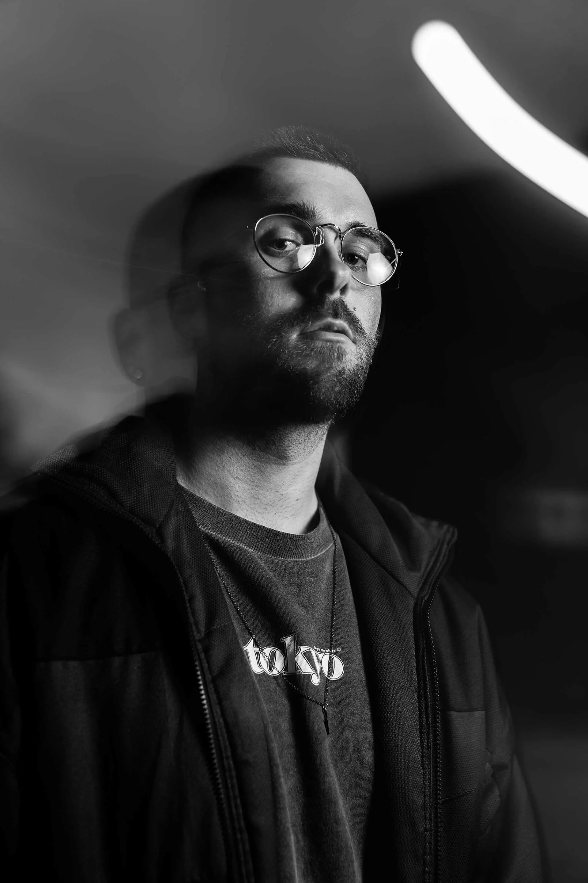 Schwarz-weiß Porträt von VAUH mit Brille, dunklem Hoodie und T-Shirt mit Tokyo-Print, porträtiert in künstlerischem Licht mit Unschärfe im Hintergrund.