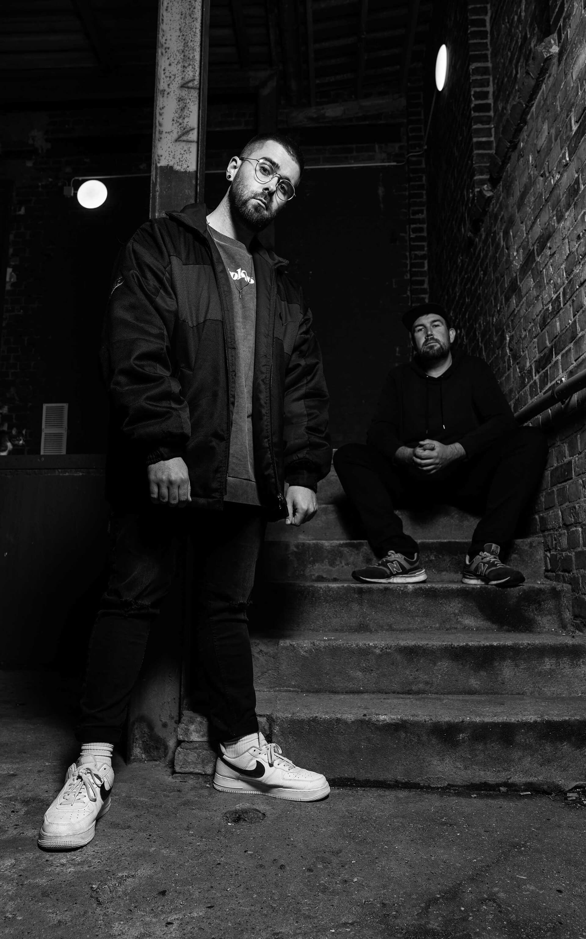 Zwei Männer in einer dunklen, urbanen Umgebung, einer steht im Vordergrund, der andere sitzt auf den Stufen im Hintergrund, beide tragen dunkle Kleidung und schauen ernst in die Kamera.