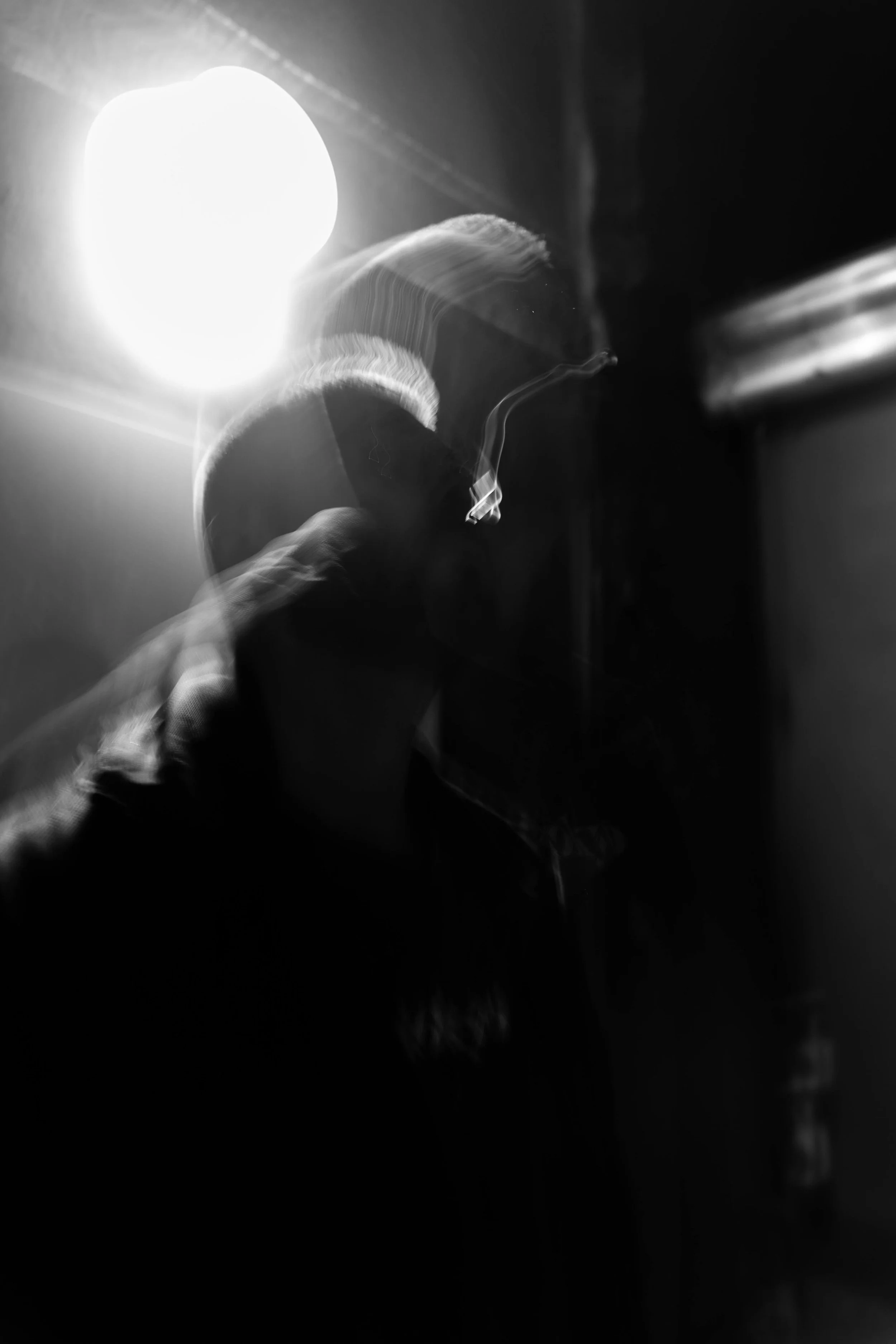 Schwarz-weiß-Fotografie VON VAUH, die eine Zigarette raucht, vor einer hellen Lichtquelle, die den Kopf der Person umgibt, mit blauer Lichtspur vom Rauch.