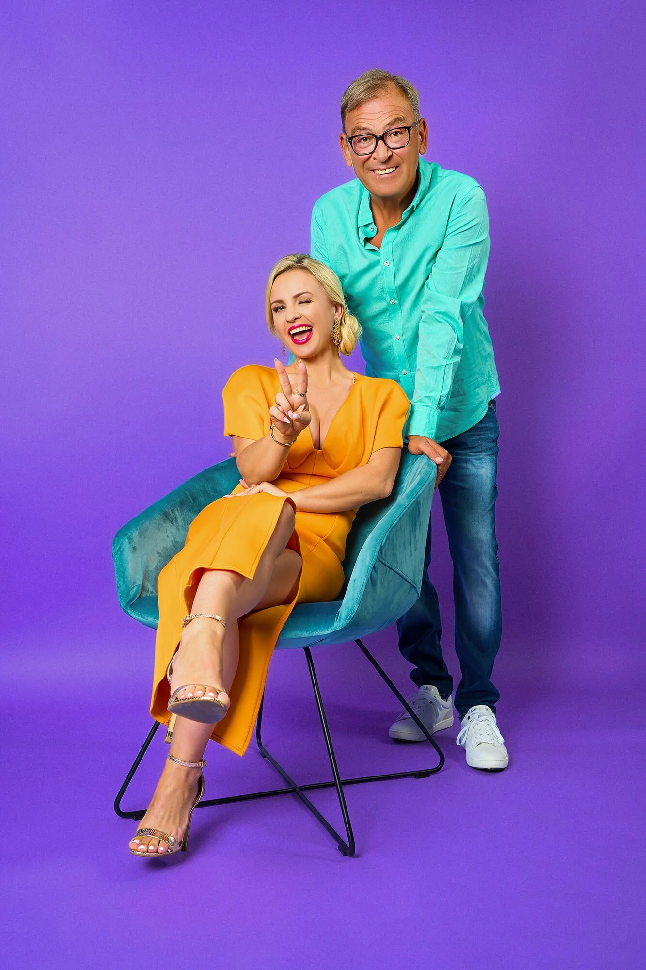 Julia Porath und Arno Müller posieren für 104.6 RTL vor einem lila Hintergrund. Julia sitzt im Vordergrund auf einem Stuhl. Arno Müller steht lächelnd dahinter.