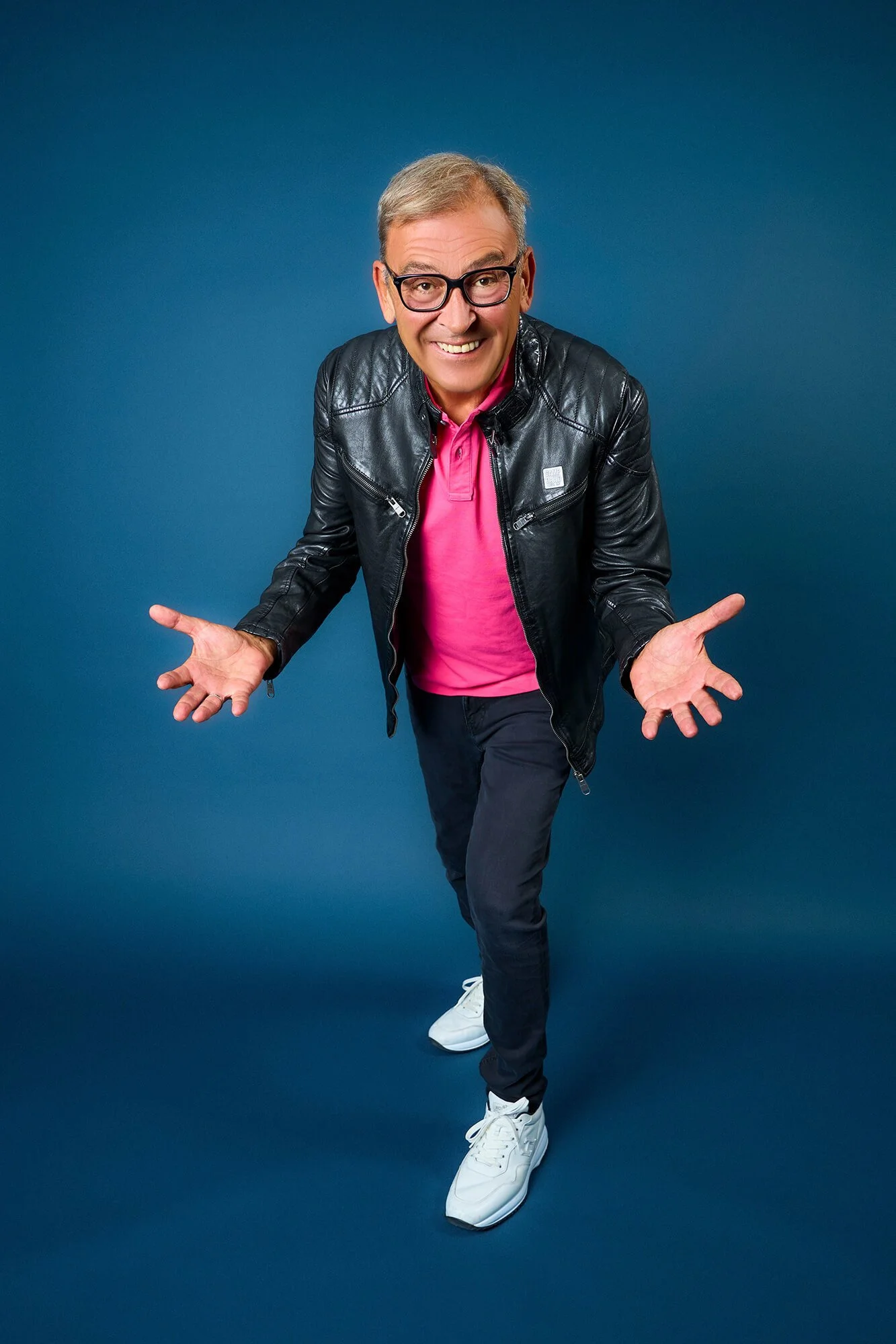 Arno Müller in schwarzer Lederjacke, pinkem Polo, Jeans, weißen Sneakers, schräg gegen die Kamera blickend, lächelnd, auf ein blaues Hintergrund