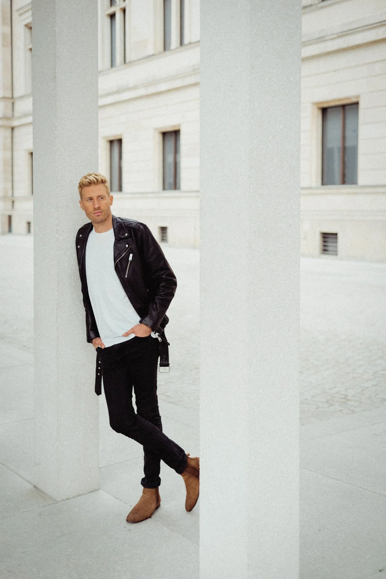 Maximilian Arland steht an einer weißen Säule vor einem historischen Gebäude, trägt eine schwarze Lederjacke, ein weißes T-Shirt, schwarze Hosen und braune Stiefel.