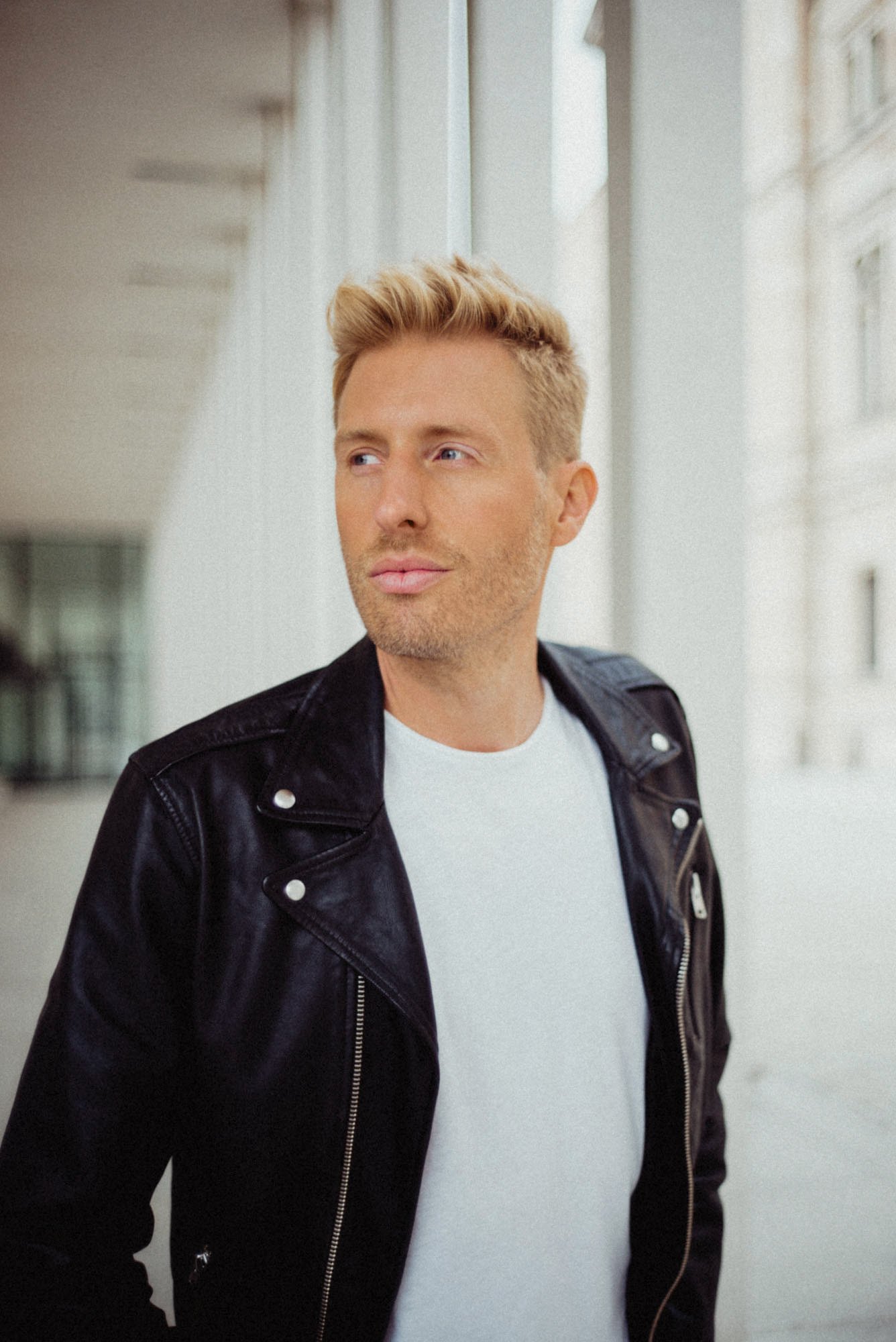 Maximilian Arland mit blondem Haar trägt ein schwarzes Lederjacke und ein weißes T-Shirt. Er steht an einer Fensterfront in einer urbanen Umgebung und schaut nach links.