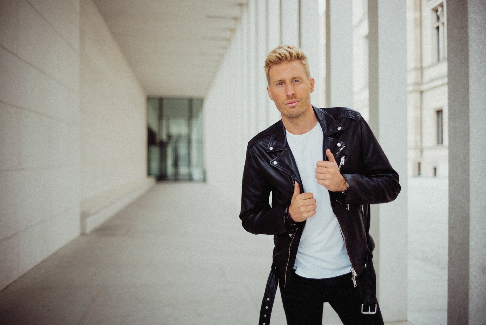 Maximilian Arland mit blonden Haaren trägt eine schwarze Lederjacke, eine weiße T-Shirt und schwarze Hosen. Er steht vor einem modernen Gebäude, schaut in die Kamera und hält die Jacke mit beiden Händen.