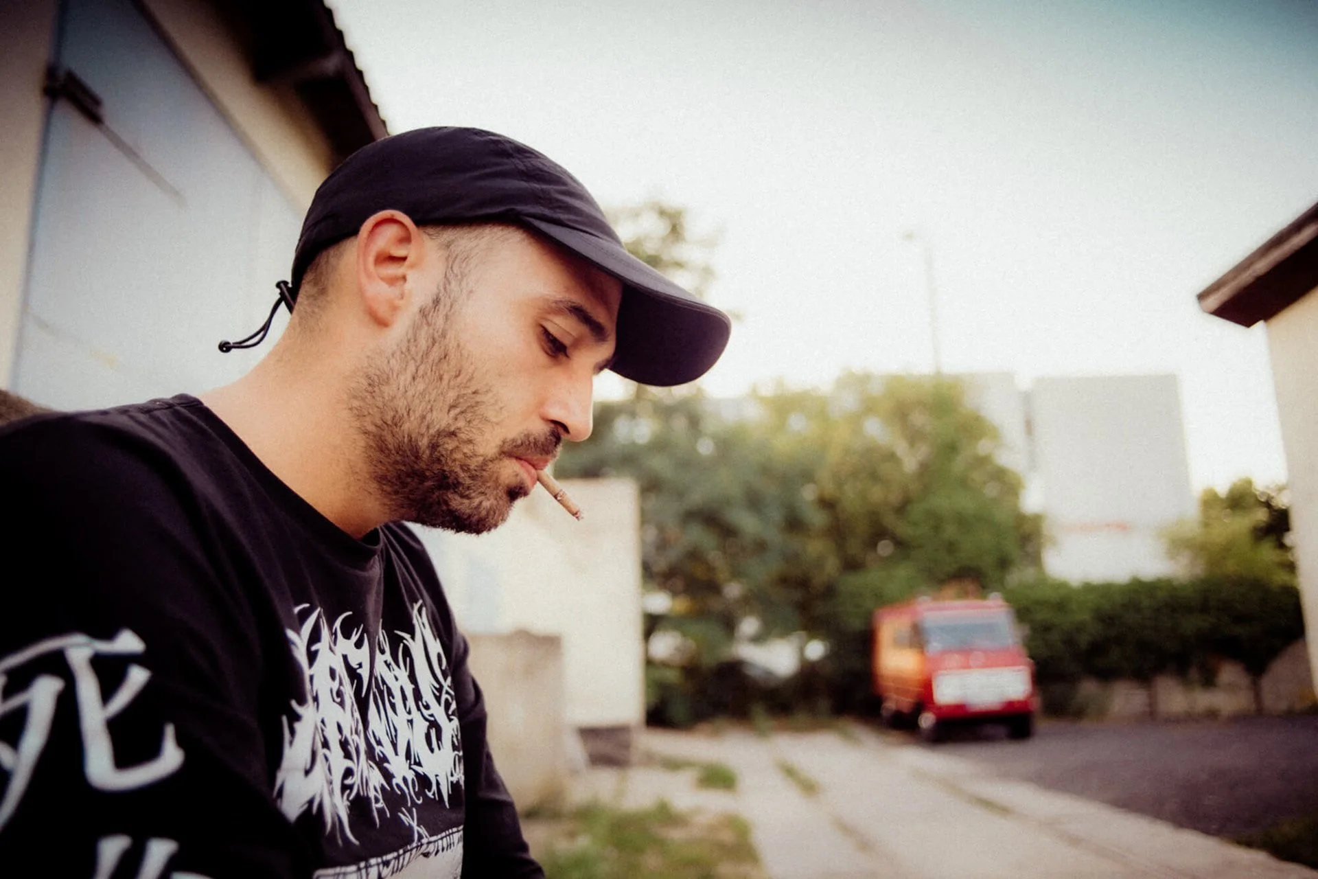 Junge Mann mit Bart, schwarzer Baseballcap und schwarzem Shirt mit weißen Mustern, der auf der Straße steht und eine Zigarette im Mund hat