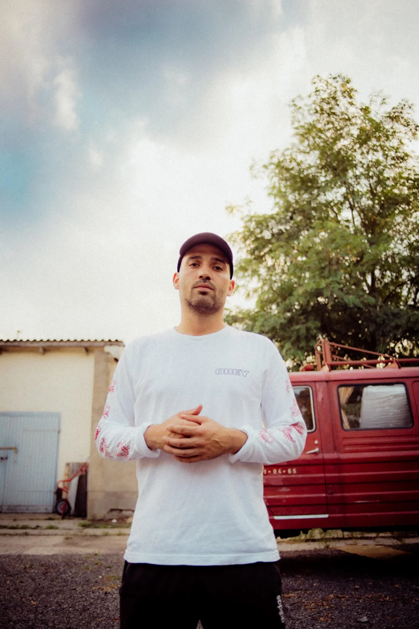 Junge Mann steht vor einem roten Fahrzeug, trägt ein weißes Oberteil mit 'OBEY'-Aufdruck, mit einer schwarzen Mütze, Blick direkt in die Kamera, im Freien bei bewölktem Himmel.