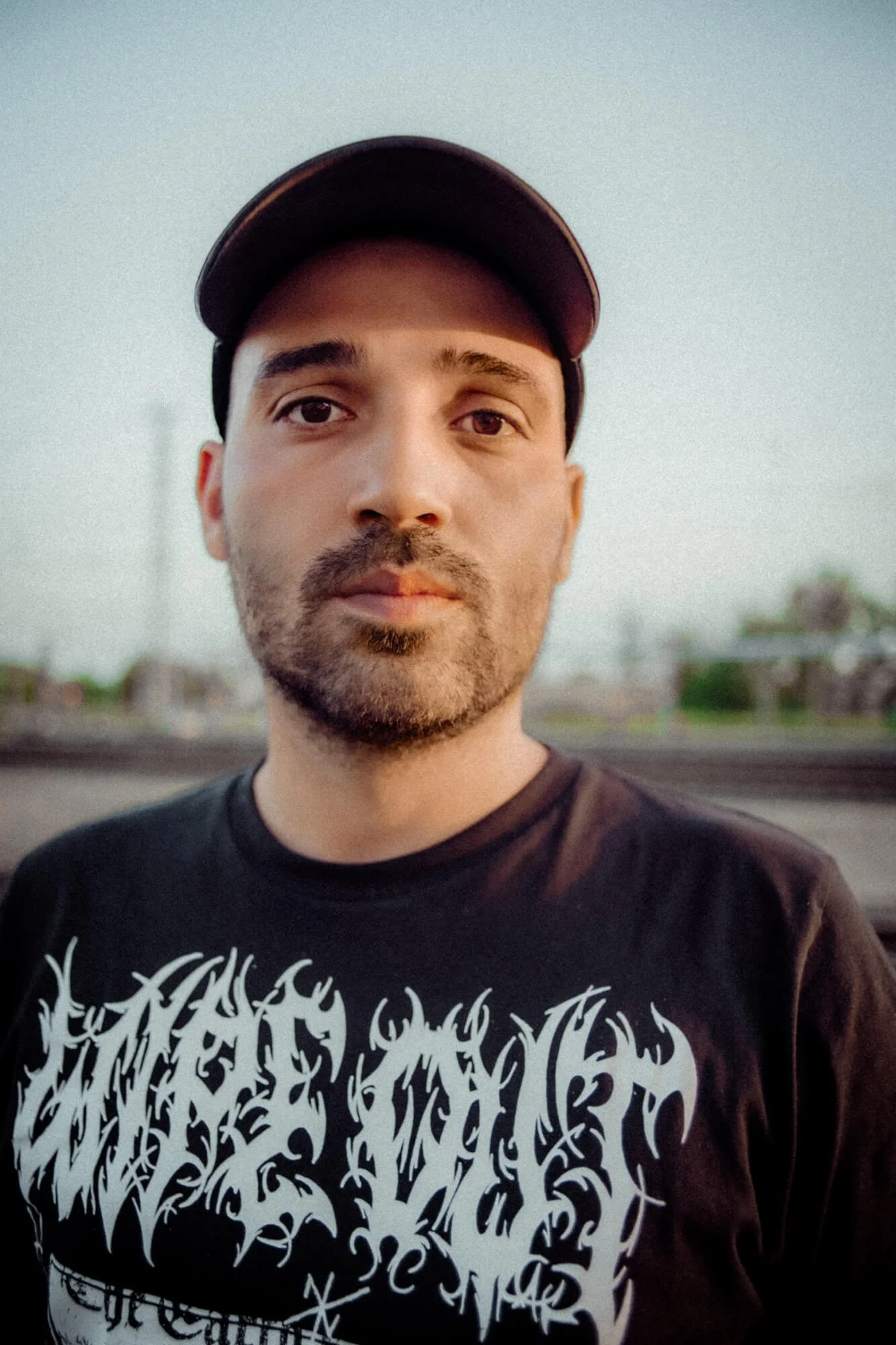 Nahaufnahme eines Mannes mit dunklem T-Shirt und schwarzen Cap, im Freien bei Sonnenuntergang, mit urbanem Hintergrund.