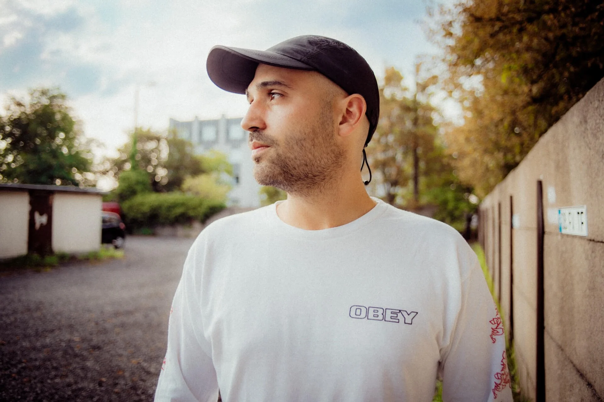 Junger Mann trägt schwarz kappe und weißen T-Shirt mit 'OBEY'-Print, sitzt neben einer Steinmauer in outdoor Umgebung, im Hintergrund Bäume und Gebäude, bewölkter Himmel