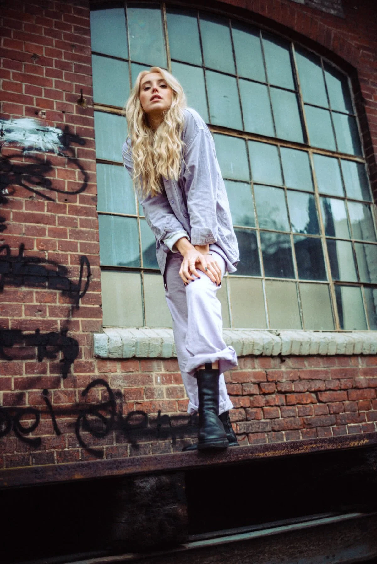 Marijke Smittenaar mit langen blonden Haaren in Latzhose und Stiefeln steht auf einem alten Eisenbahnwagen vor einer Hauswand mit großen Fenster und Graffiti.