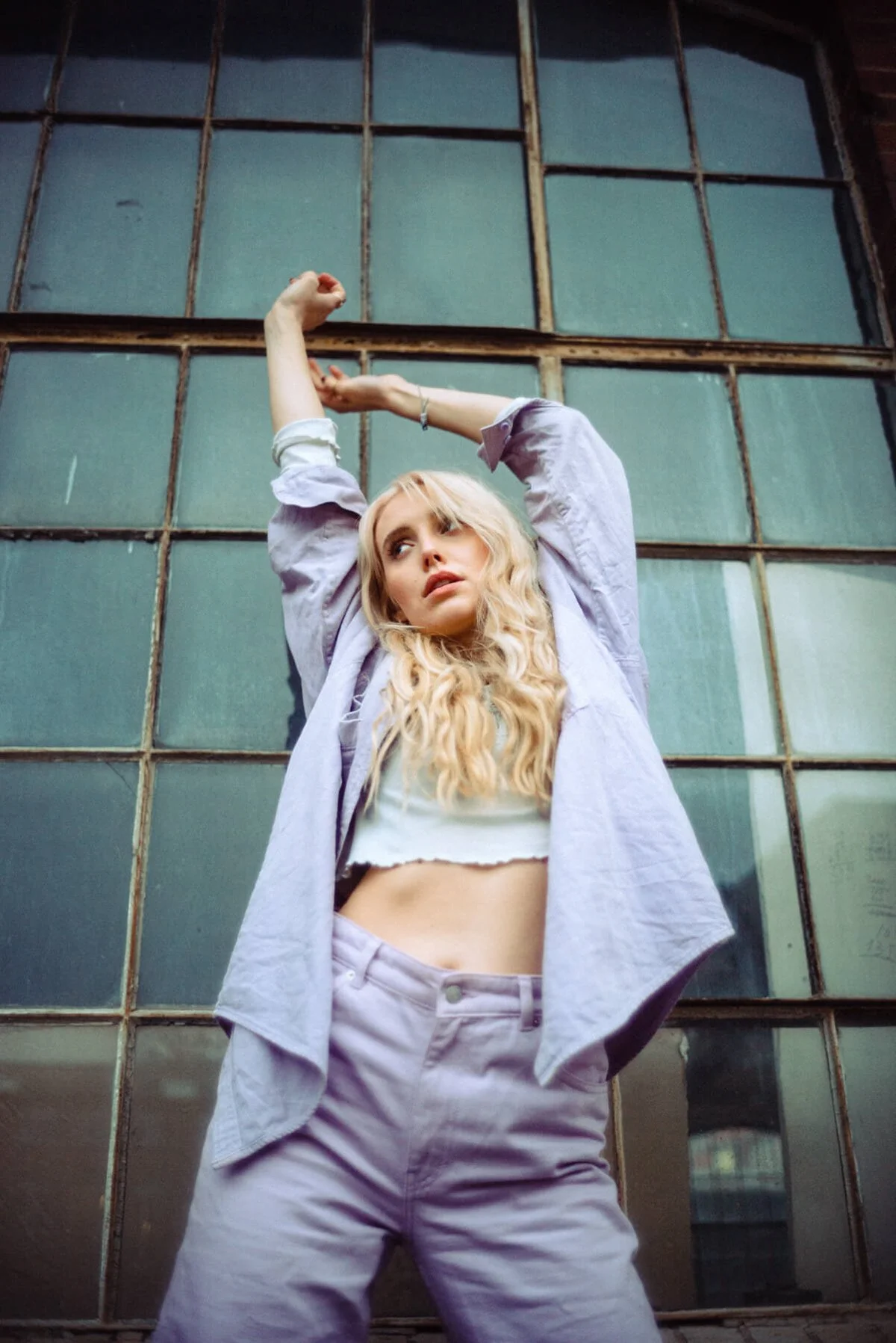 Marijke Smittenaar mit blonden Locken pose vor einer alten, großen Fensterwand, trägt eine offene, helle Jacke, ein Cropped-Top und Hosen, Arme hoch, blickt nach links.