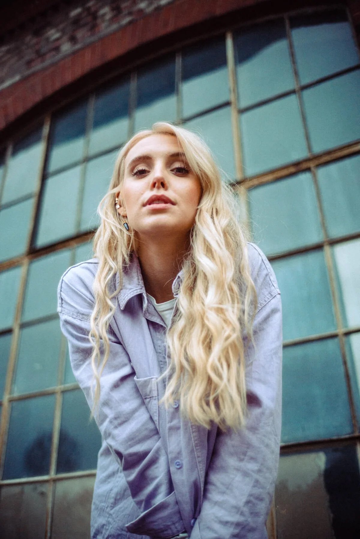 Marijke Smittenaar mit langen, lockigen blonden Haaren, trägt eine graue Jacke und steht vor einem alten Gebäude mit großen Glasfenstern.