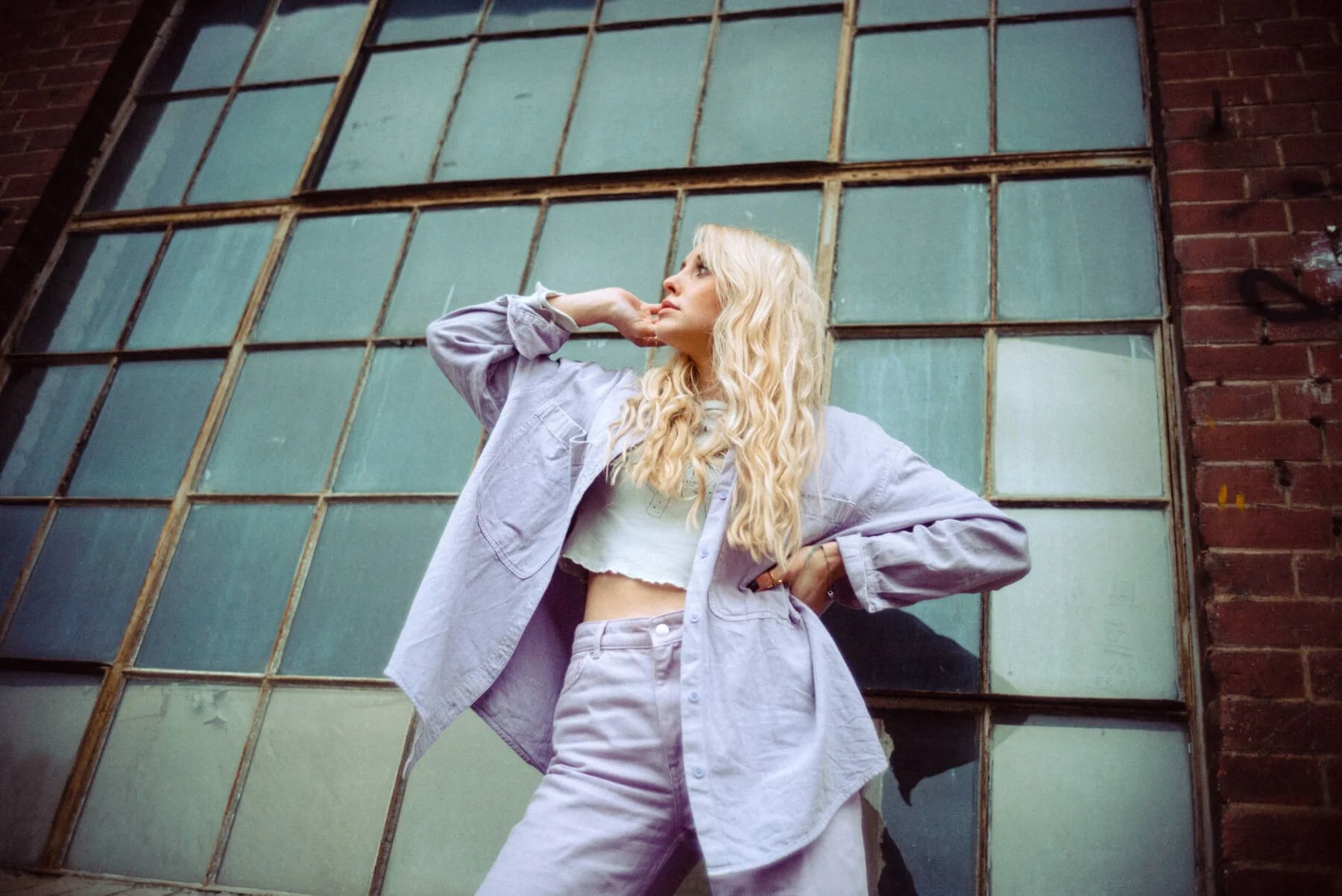 Marijke Smittenaar mit blonden, lockigen Haaren posiert vor einem alten, großen Fenster mit quadratischen Glasfeldern in einer Lagerhalle oder Fabrik.