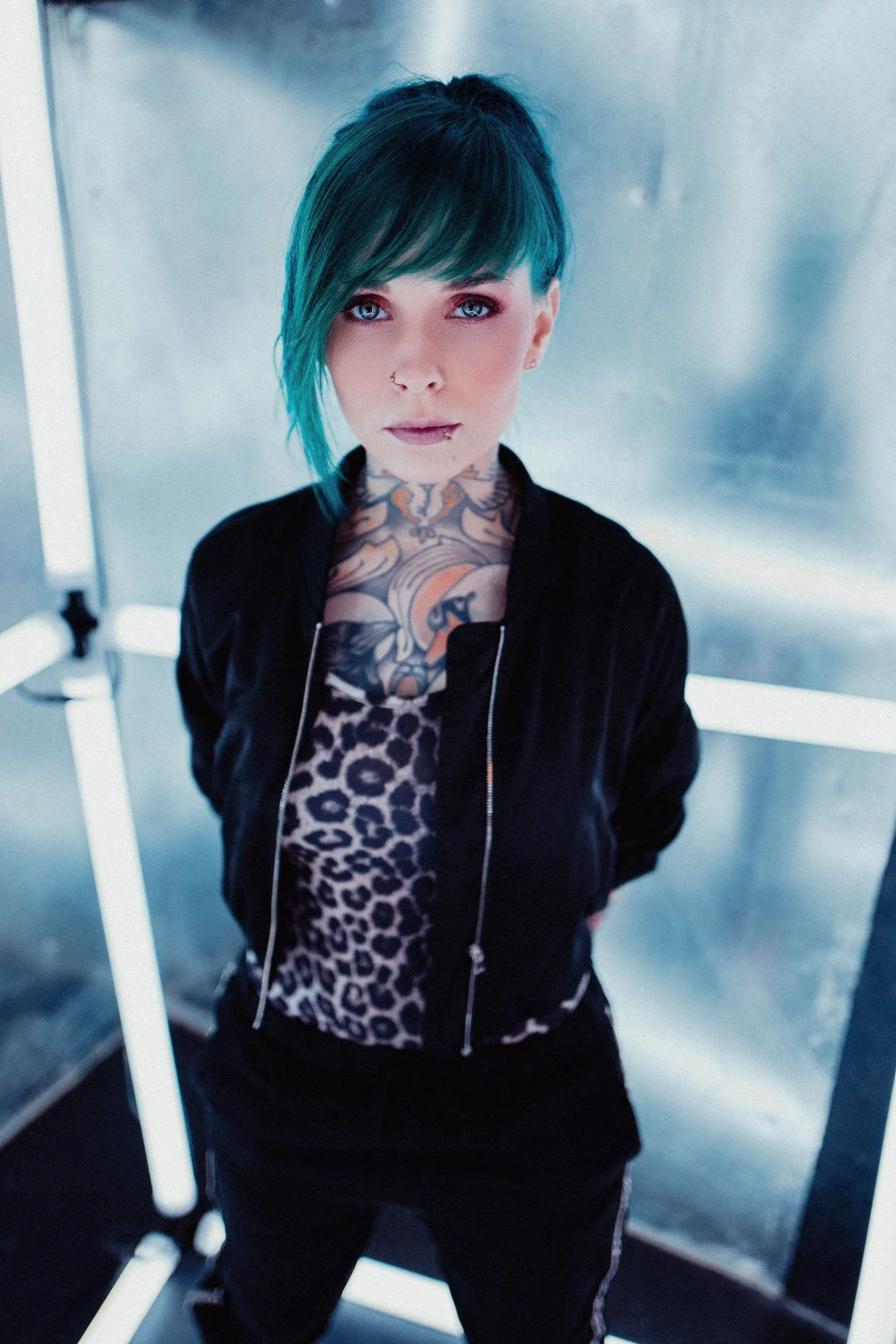 Jazzy Gudd mit kurzen blauen Haaren, Tattoos im Hals- und Brustbereich, schwarz-weißem Leopardenmuster-Shirt und schwarzer Jacke, steht vor einer metallischen Wand in Neonbeleuchtung.