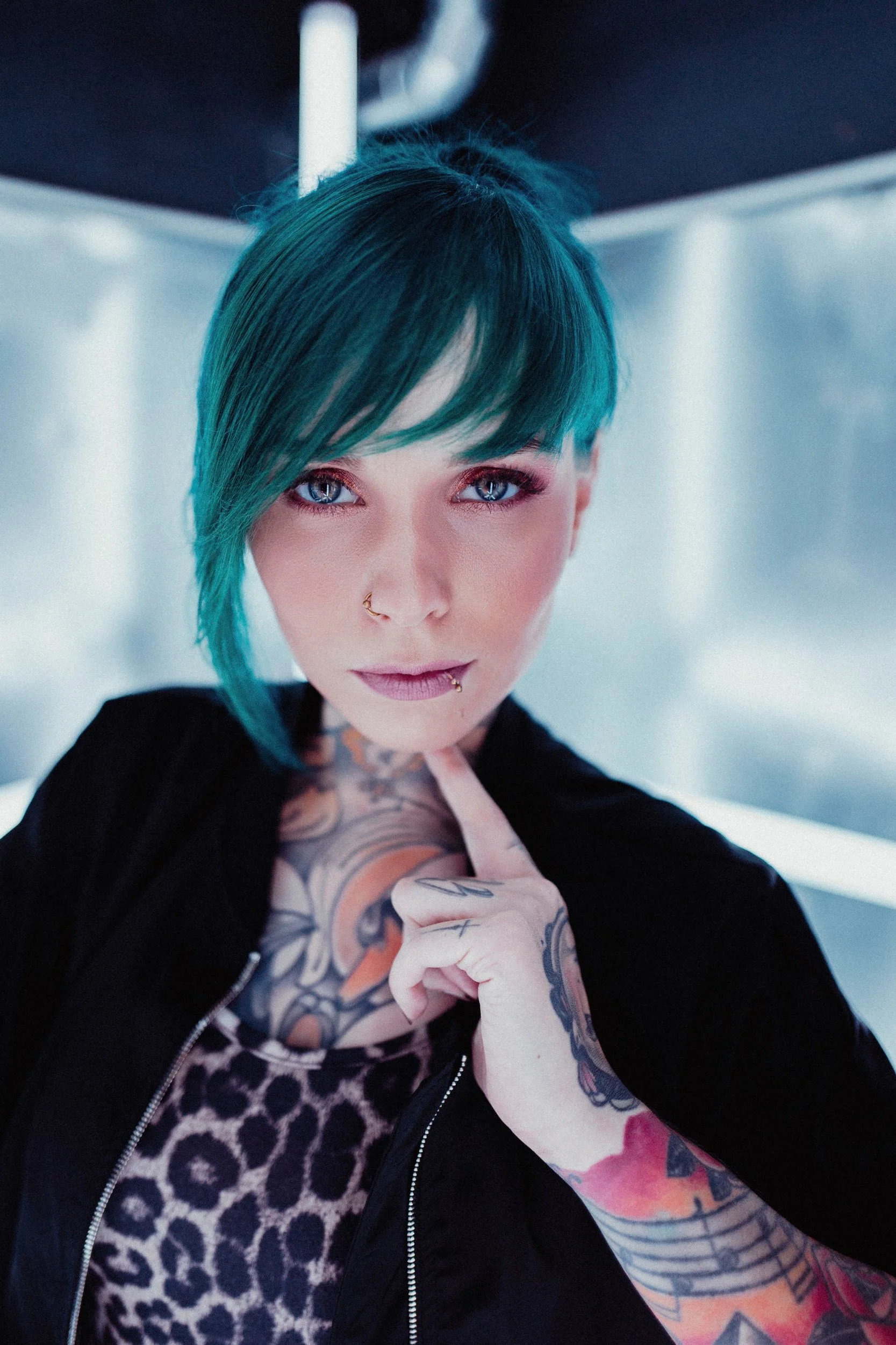 Nahaufnahme von Jazzy Gudd mit blauer Kurzhaarfrisur, tätowiertem Hals und Armen, trägt dunkle Kleidung und hat Piercings im Gesicht, blickt direkt in die Kamera.