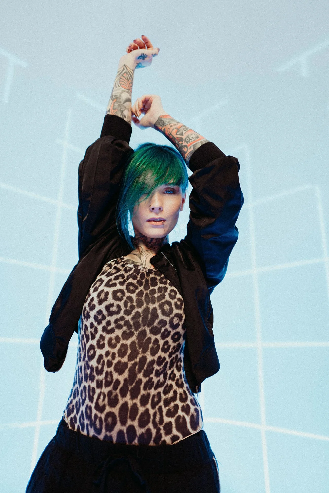 Jazzy Gudd mit grünen und blauen Haaren, tattoos, animal print Top und schwarzer Jacke, in einem Raum mit blauer Beleuchtung und Gittermuster an der Wand.