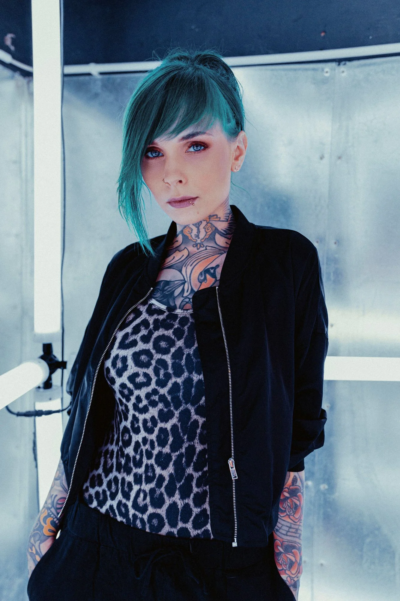Jazzy Gudd mit orangem, blauen und schwarzen Haaren, Tattoos, schwarzen Jacke und Leoparden-Top vor einer metallischen Wand im Neonlicht.