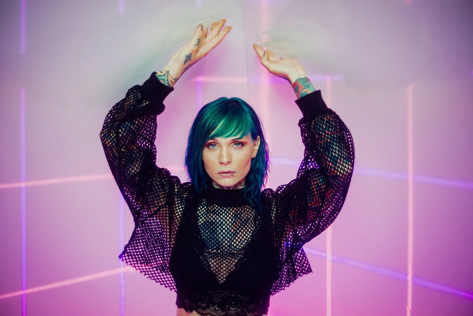 Jazzy Gudd mit blauer und grüner Haarfarbe in Netzoberteil posiert vor einer pinken Wand mit Neonlinien.