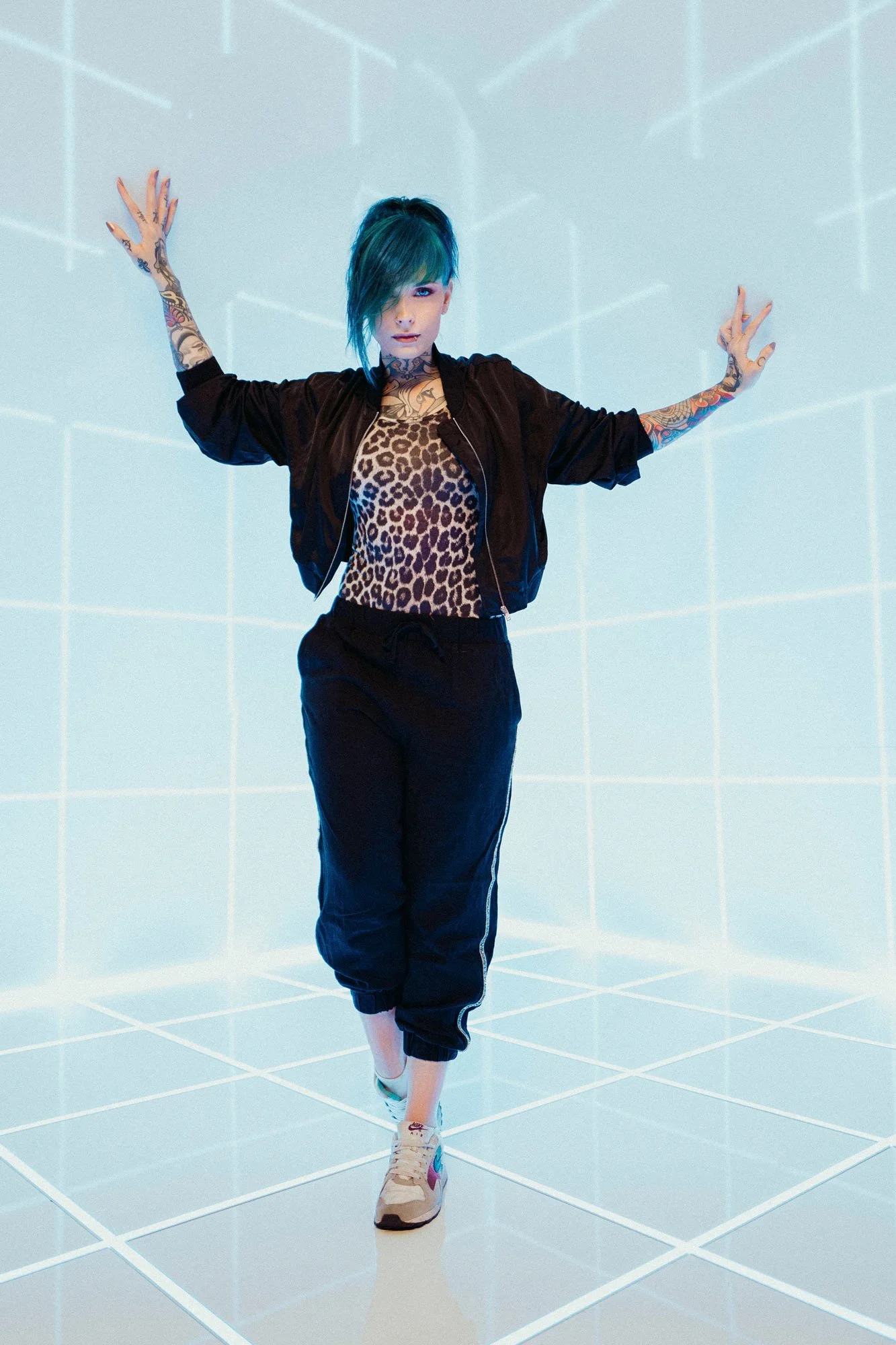 Jazzy Gudd mit blauer Frisur in einem futuristischen Raum posiert. Sie trägt eine Leoparden-Top, eine schwarze Jacke und Jogginghosen.