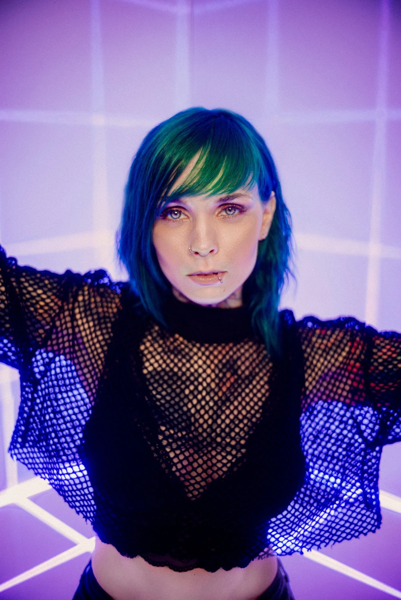 Jazzy Gudd mit grünen und blauen Haaren in einem schwarzen Netzshirt, in Neonlicht getaucht.