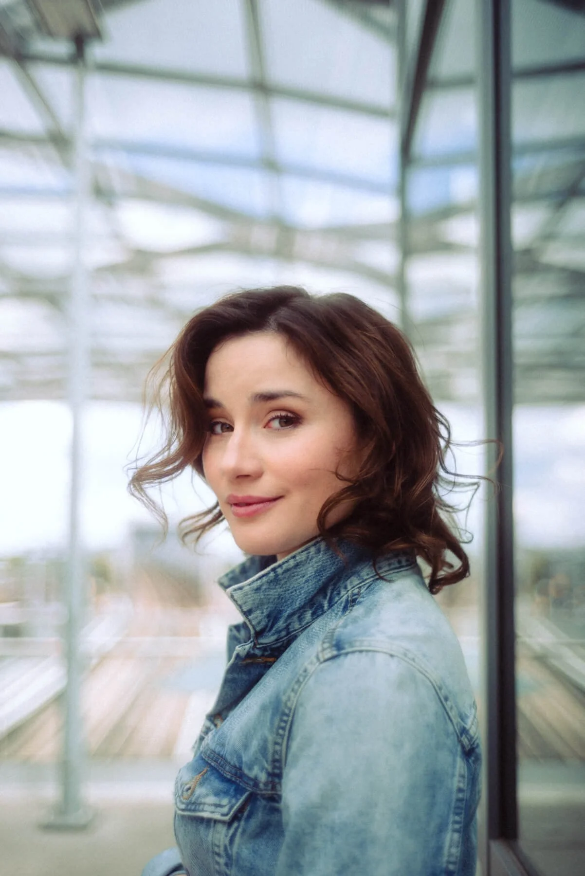 Jeannine Michèle Wacker mit braunen, lockigen Haaren trägt eine Denim-Jacke und blickt lachend in die Kamera, im Hintergrund ist eine moderne Glasstruktur sichtbar.