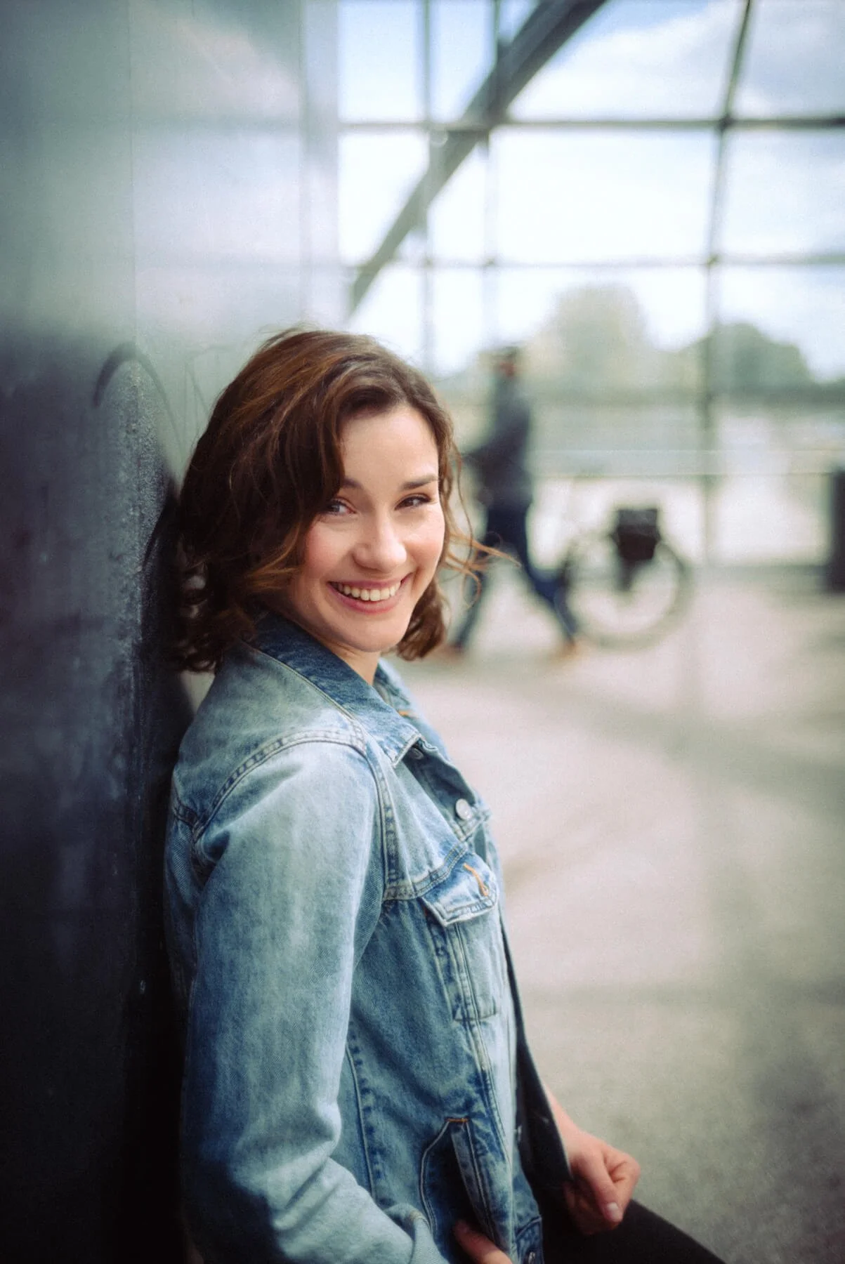 Jeannine Michèle Wacker mit braunen, lockigen Haaren, das in einem Freiluftbahnhof gegen eine Wand lehnt und lächelt.